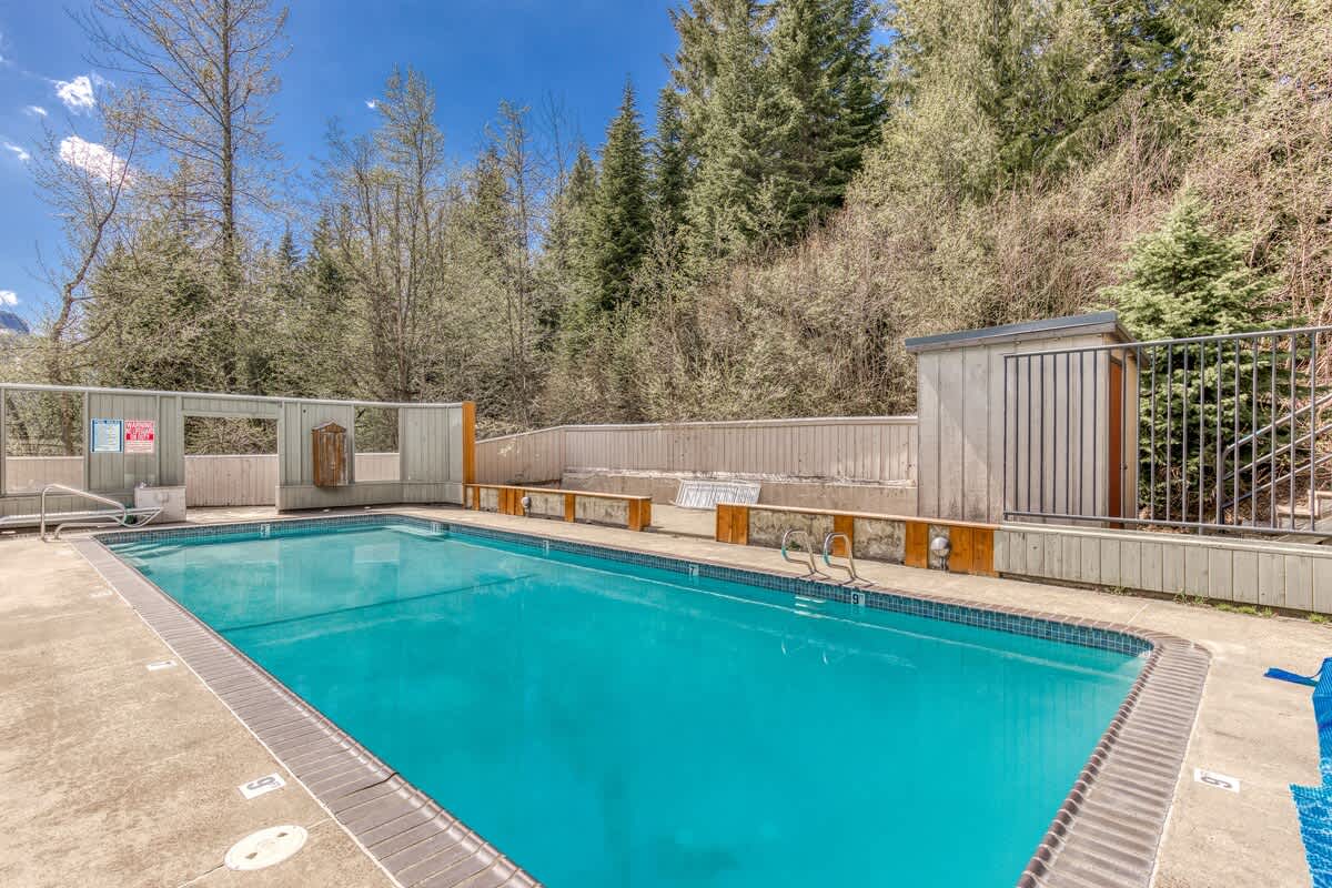 An outdoor rectangular swimming pool with blue water, surrounded by a concrete deck and a low fence, with tall evergreen trees and a mountain visible in the background under a bright blue sky. Benches are along one side of the pool.