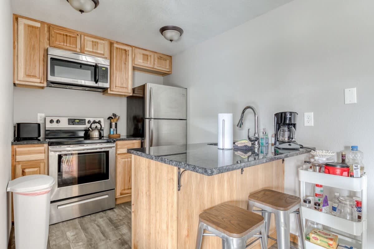Compact kitchen equipped with stainless steel appliances including a stove, microwave, and refrigerator, light wood cabinetry, dark granite countertops, and a small island with two bar stools.