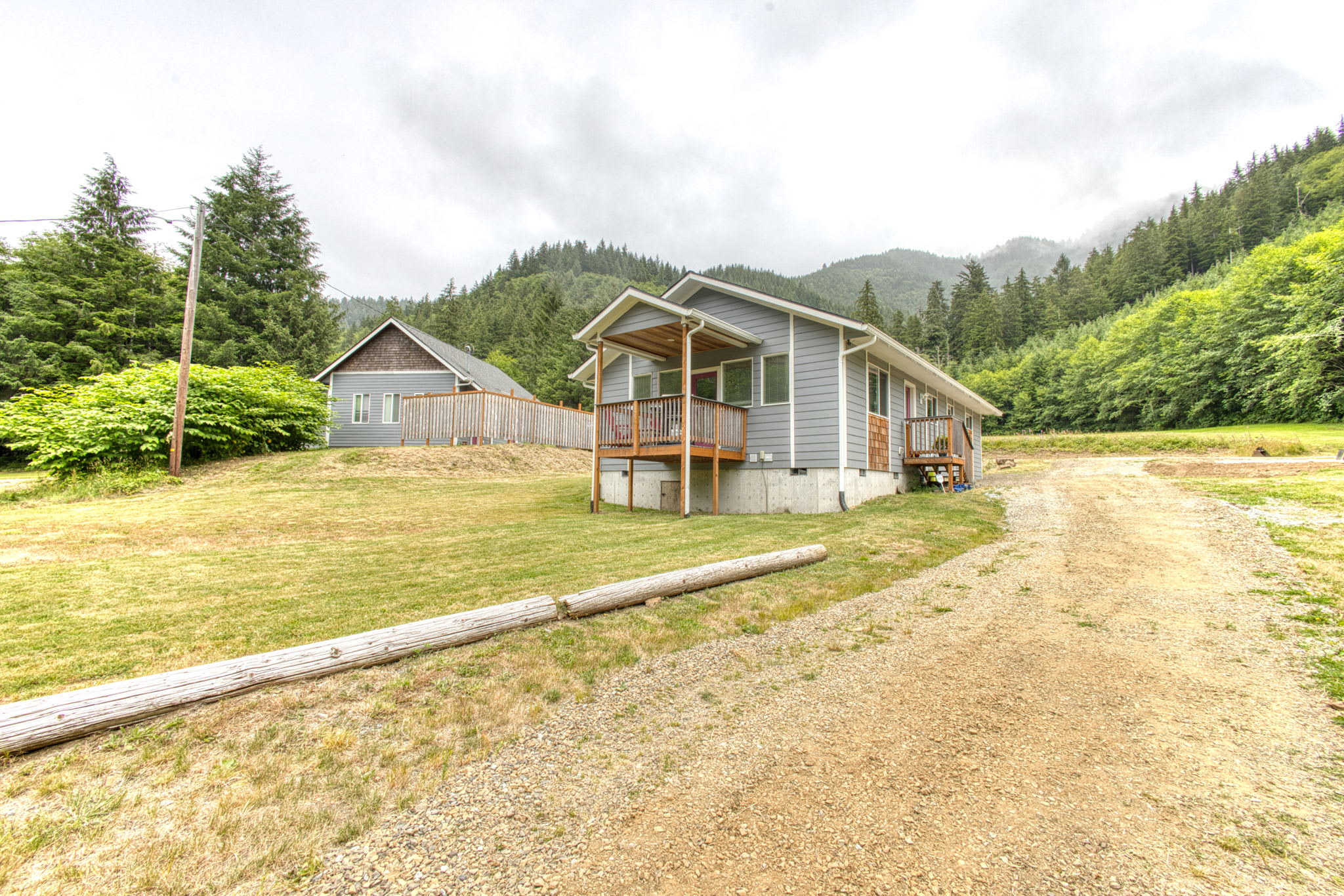 A side view of two light gray houses, one with a covered porch, are situated on a grassy and gravel landscape. A dirt driveway leads past the houses, with dense green forests and misty mountains in the background.