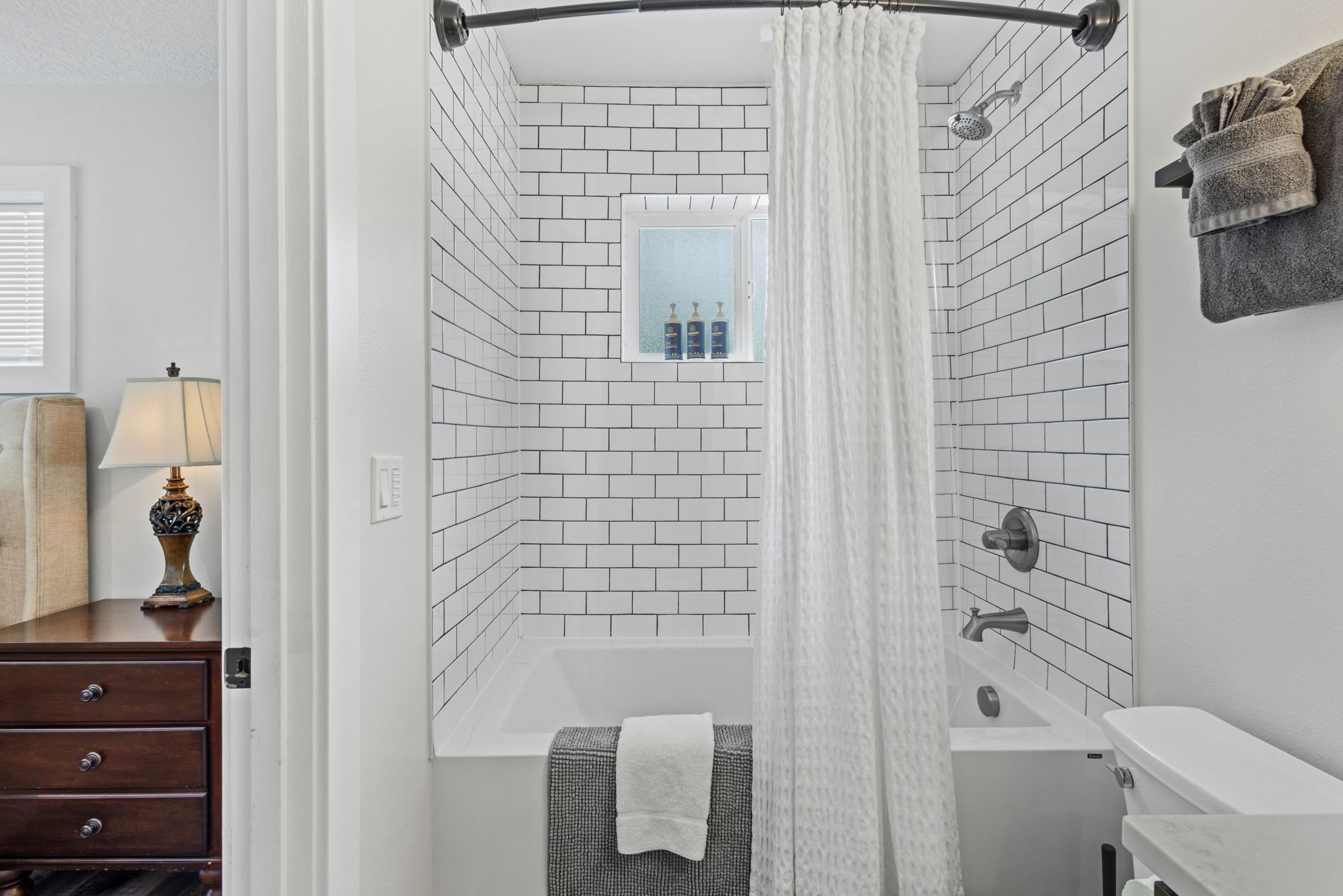A bright bathroom featuring a white bathtub enclosed by white subway tiles with dark grout, a white waffle-textured shower curtain, and a small window above the tub displaying toiletries.