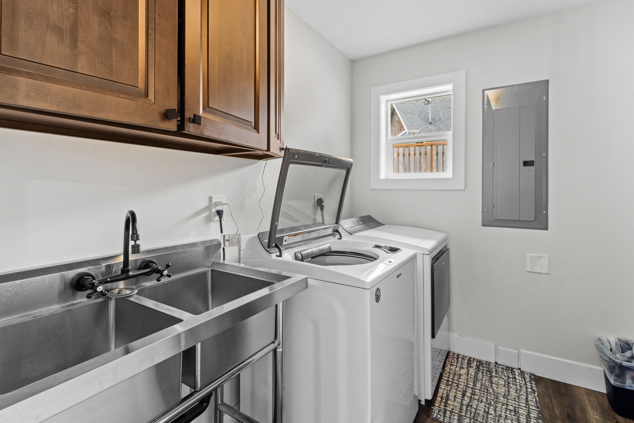 A functional laundry room equipped with a top-loading washing machine, a dryer, and a stainless steel utility sink with a black faucet. Dark wood cabinets are mounted above the sink, and a window provides natural light.
