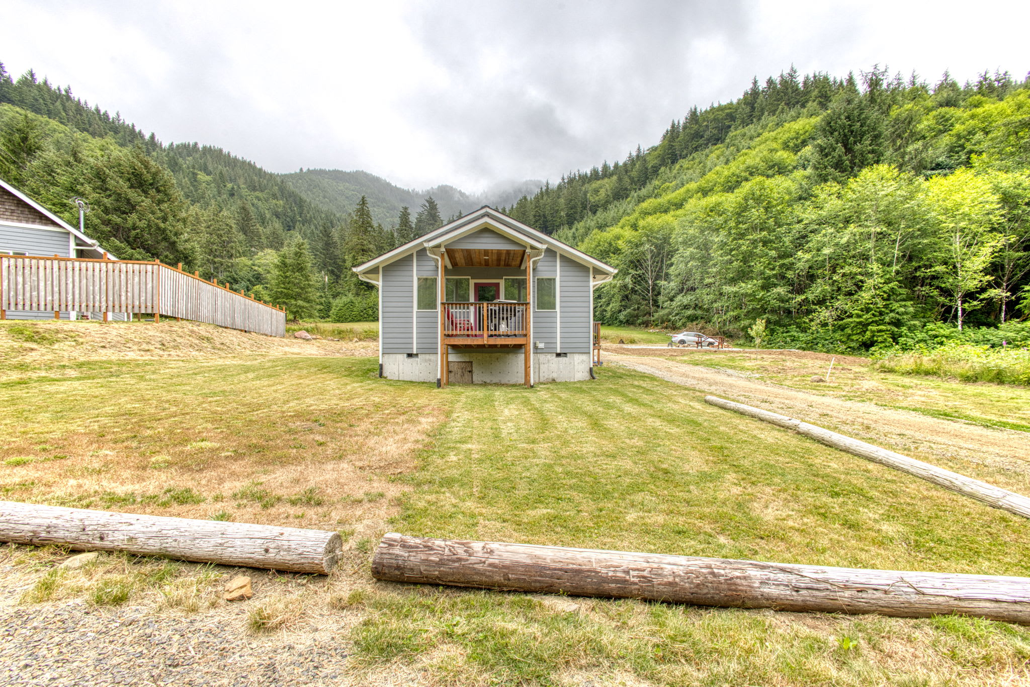The front exterior of a light gray house with a covered porch and wooden railing is visible, set on a grassy lawn with a gravel path. Dense green forests and misty mountains form the backdrop under a cloudy sky.