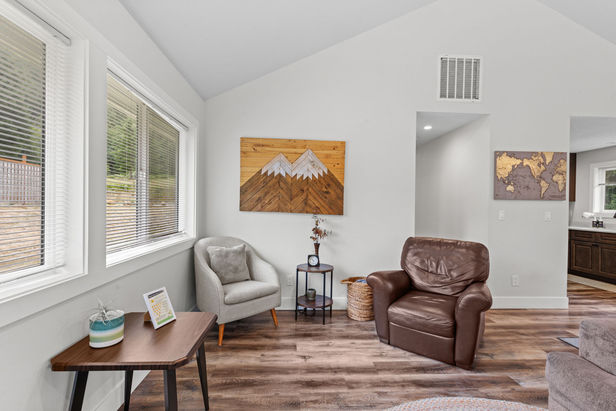 Bright corner of the living room with a light grey armchair and a brown leather armchair, separated by a small side table, under a wooden mountain-themed wall art piece, with large windows providing natural light.