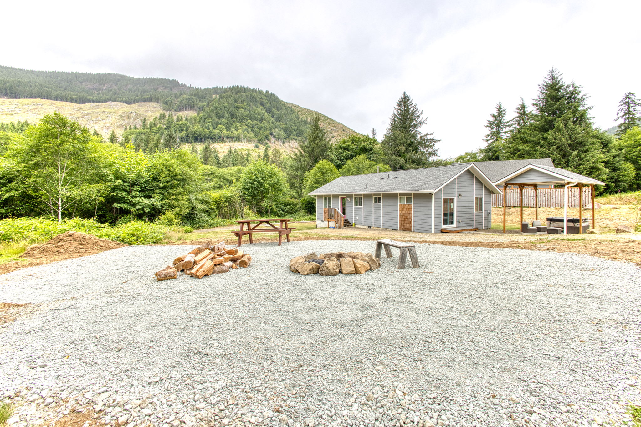 A spacious gravel area features a stone fire pit with wooden benches and a picnic table, surrounded by stacked firewood. A light gray house is visible in the background, with lush green trees and a distant mountain.