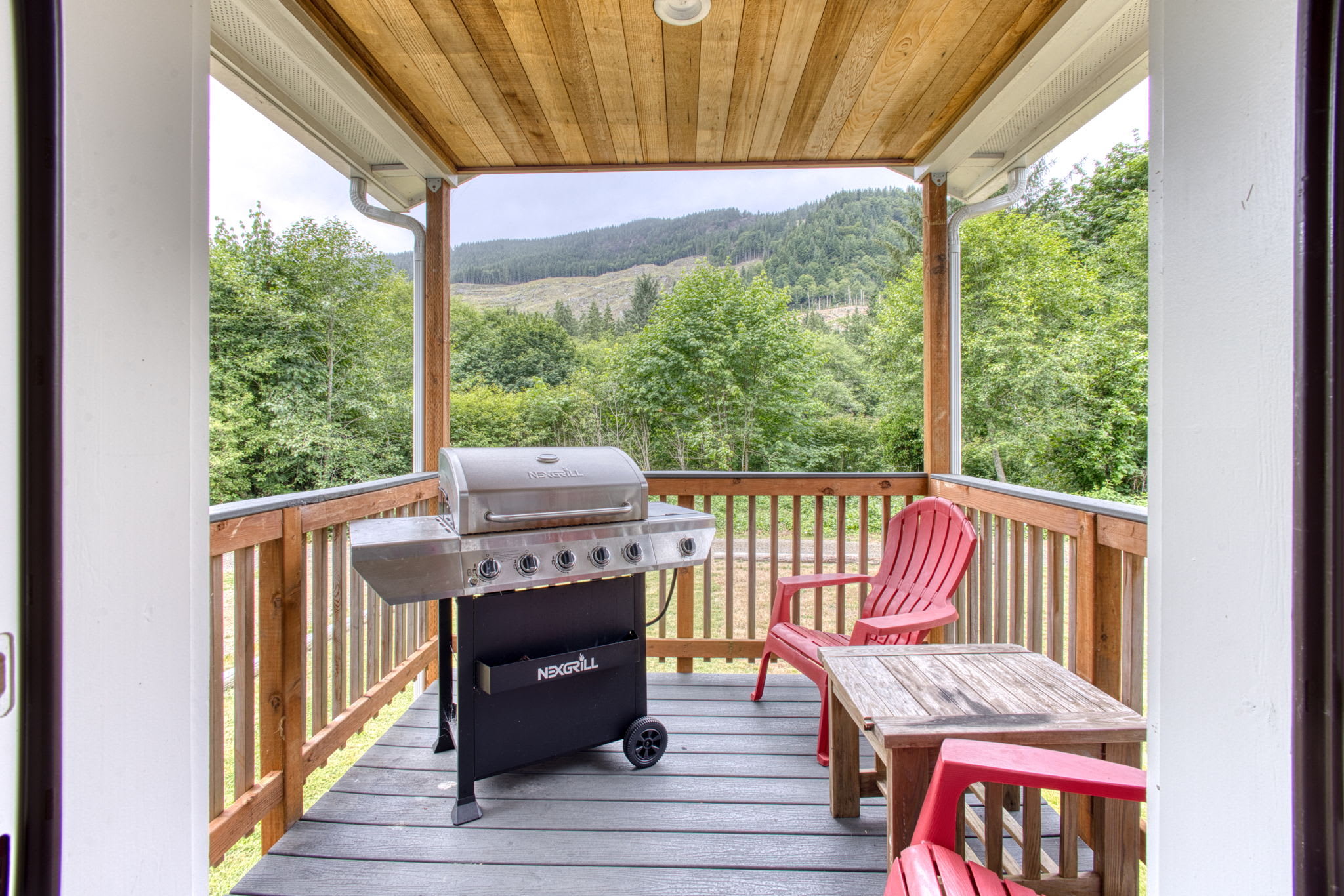 Covered outdoor deck with a gas grill and two red Adirondack chairs next to a small wooden table, offering views of lush green forested hills in the distance.