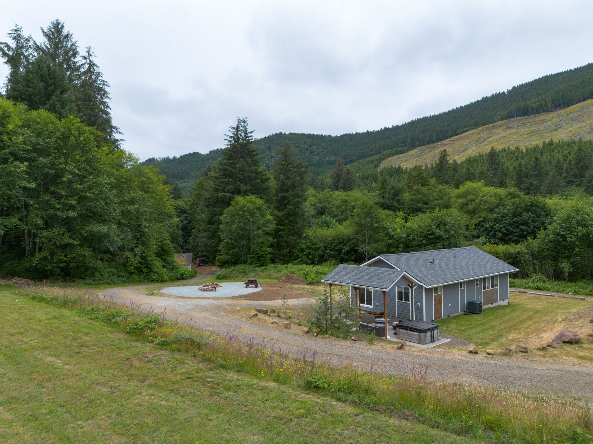 An aerial view of a gray house with a dark gray roof, situated on a gravel driveway amidst a lush green landscape with dense forests and mountains in the background under a cloudy sky. A fire pit area is visible in the foreground.