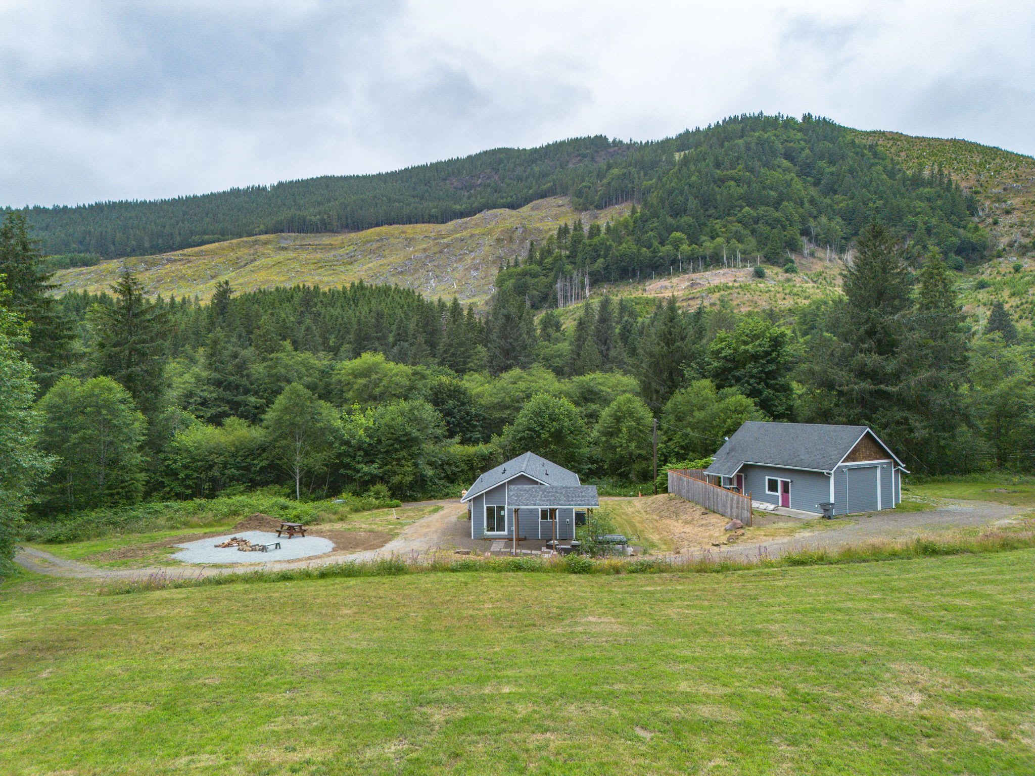 An aerial view shows two light gray houses on a property with a gravel driveway, surrounded by green grass and dense forests. A large cleared area on the distant hillside suggests logging activity.