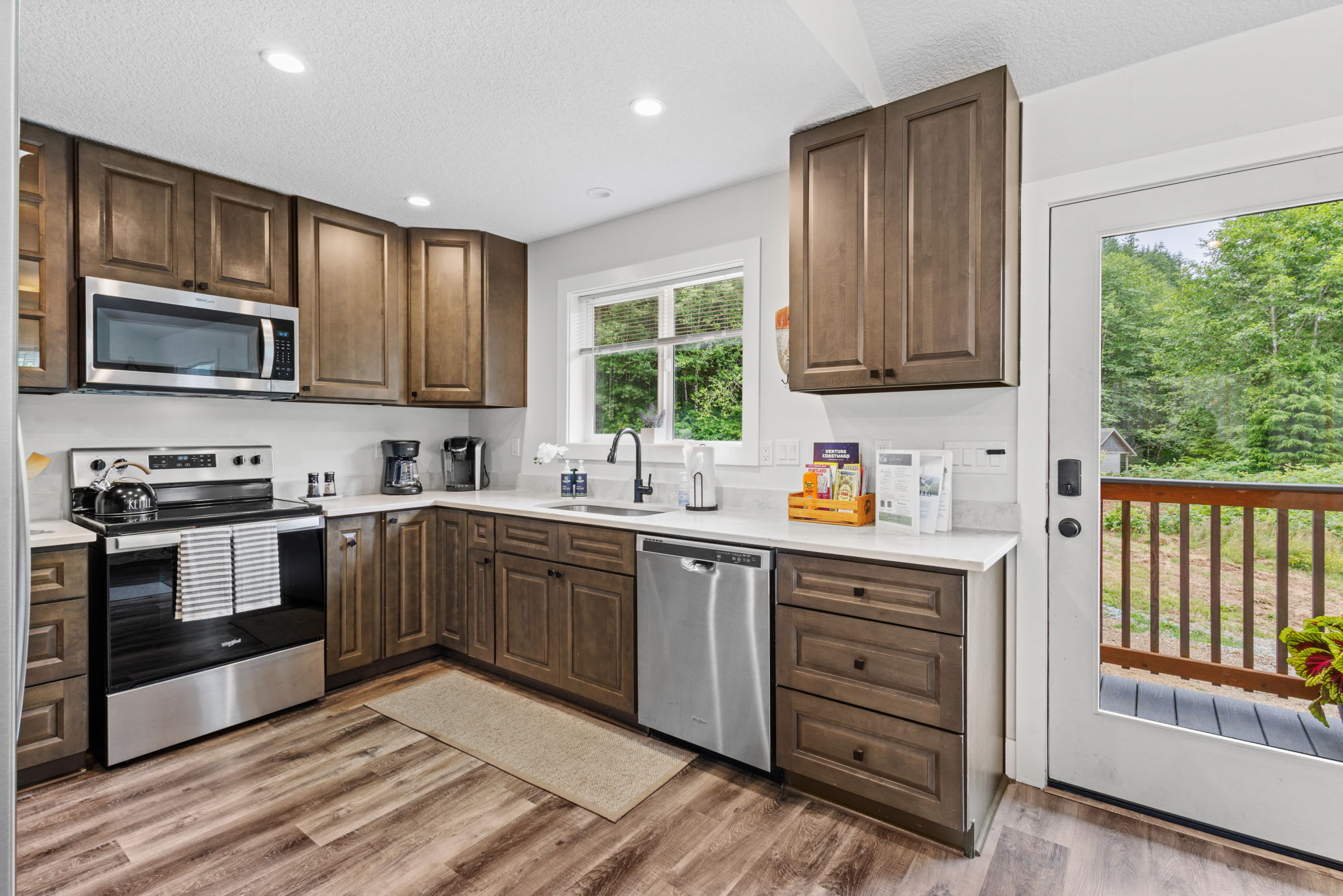 Well-equipped kitchen with dark wood cabinets, white quartz countertops, stainless steel appliances, and a window above the sink, with a white door leading to an outdoor deck.