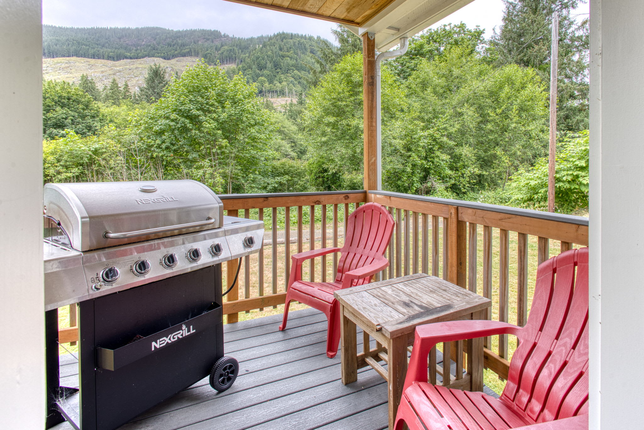 A cozy covered porch features a stainless steel Nexgrill BBQ, two bright red Adirondack chairs, and a small wooden side table, all set against a backdrop of lush green trees and distant hills.