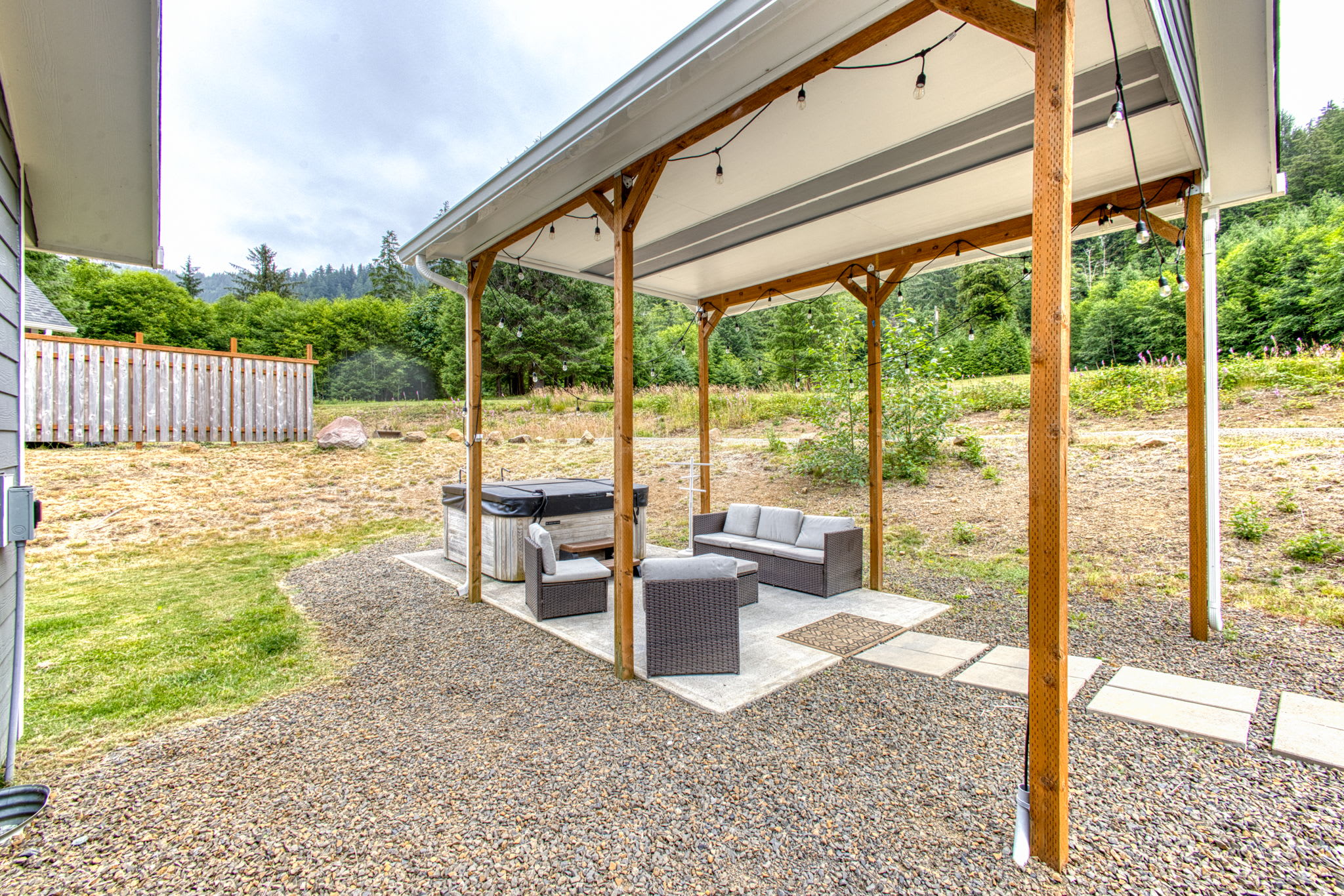 A covered outdoor area features a dark hot tub next to wicker outdoor seating with light cushions, all on a concrete pad with string lights overhead. The surrounding area is gravel and grass, with a wooden fence and forest in the background.
