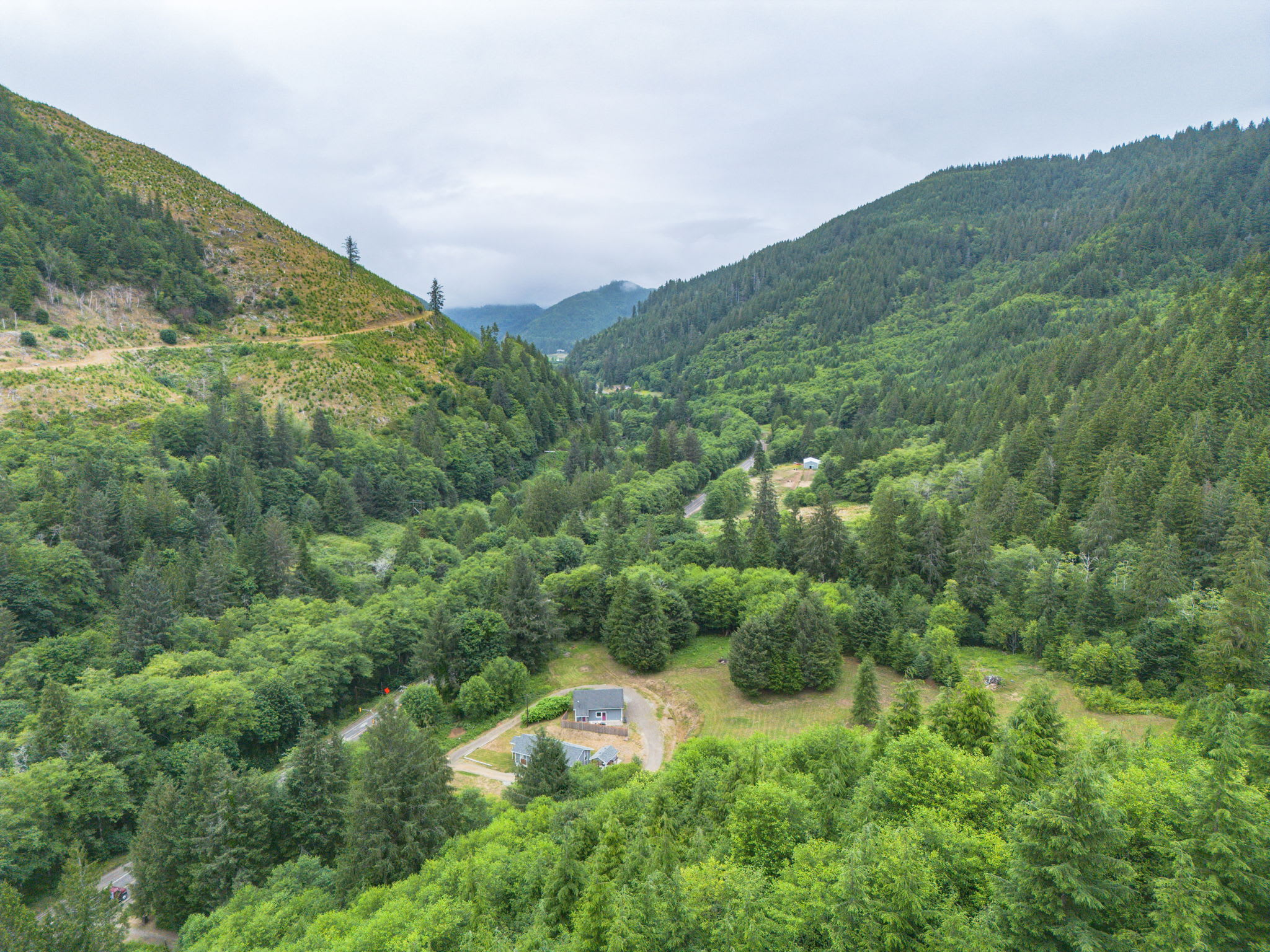 A wide aerial shot showcasing a house and its surroundings within a vast, forested valley with mountains under a cloudy sky. The property features a gravel driveway and is surrounded by lush green trees.