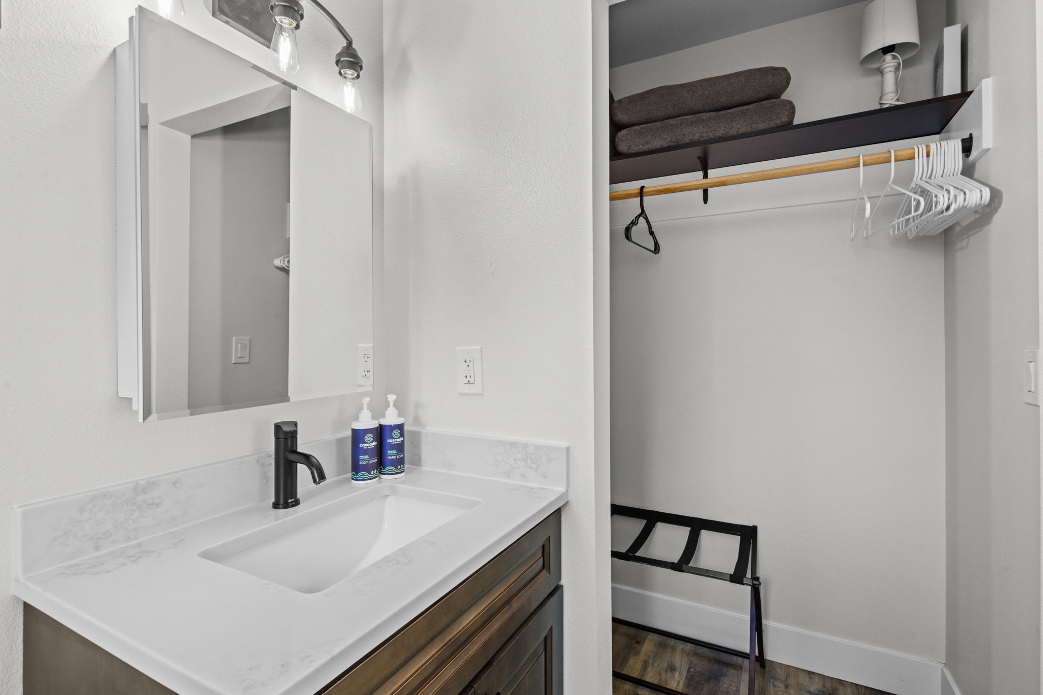 A close-up of the bathroom vanity with a white quartz countertop, rectangular sink, black faucet, and two bottles of soap. A mirrored medicine cabinet is above the sink, and an open closet with hangers and folded towels is visible to the right.