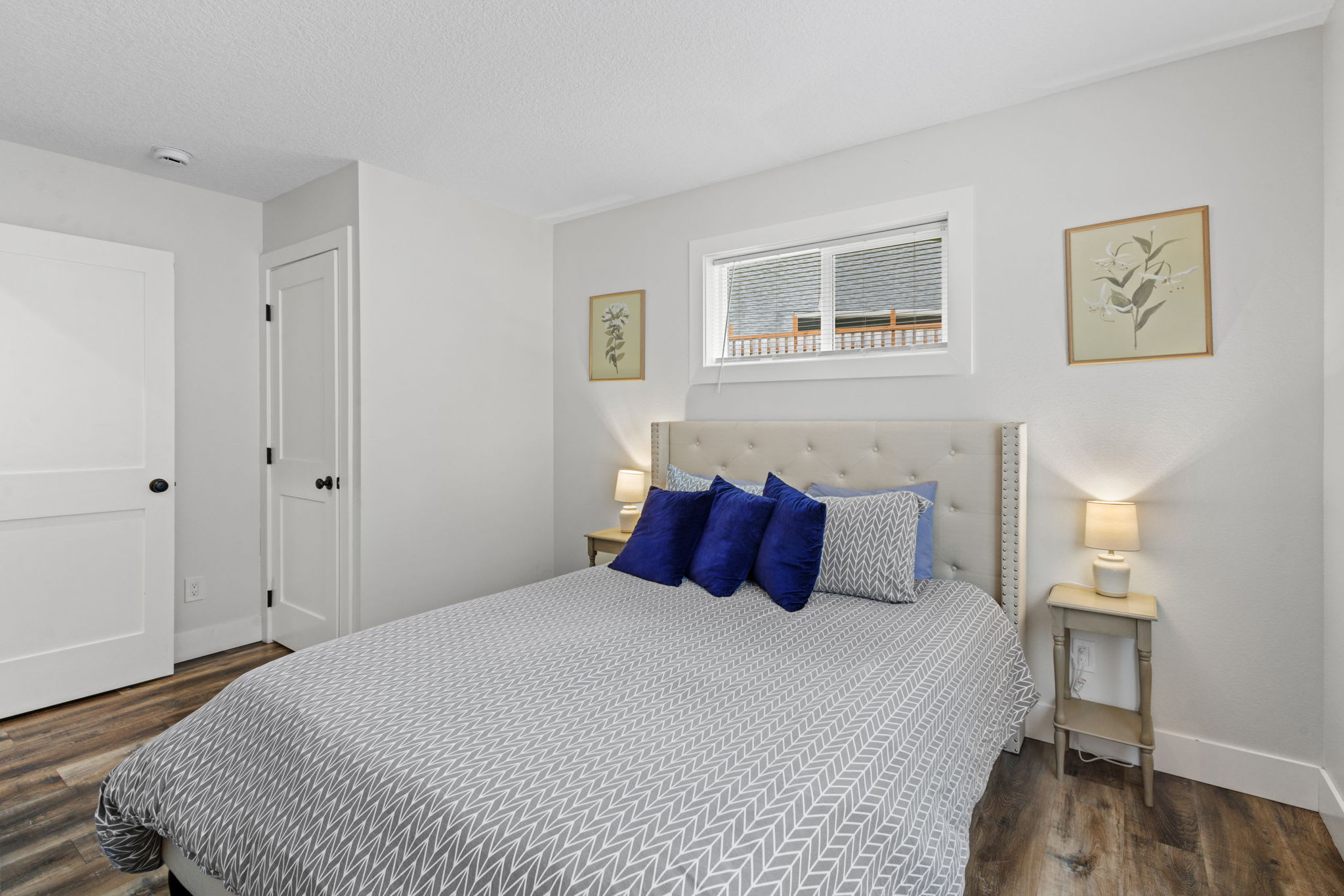 A cozy bedroom featuring a queen-sized bed with a light gray tufted headboard, blue and gray pillows, and a gray patterned duvet. Two small light-colored nightstands with lamps flank the bed, and two white doors are visible to the left.