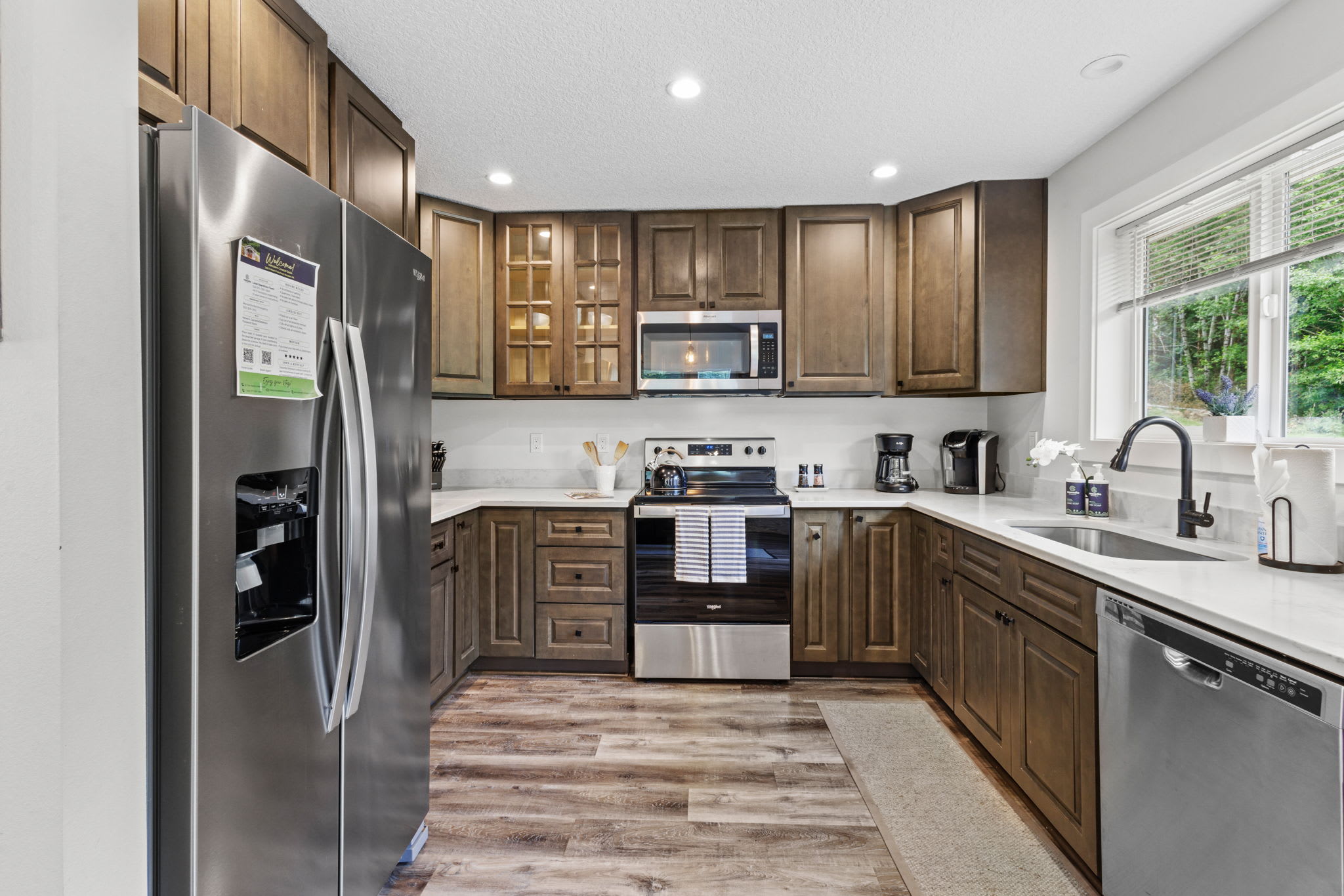 Modern kitchen with dark wood shaker-style cabinets, white quartz countertops, stainless steel appliances including a refrigerator, range, and microwave, and light wood-look flooring.