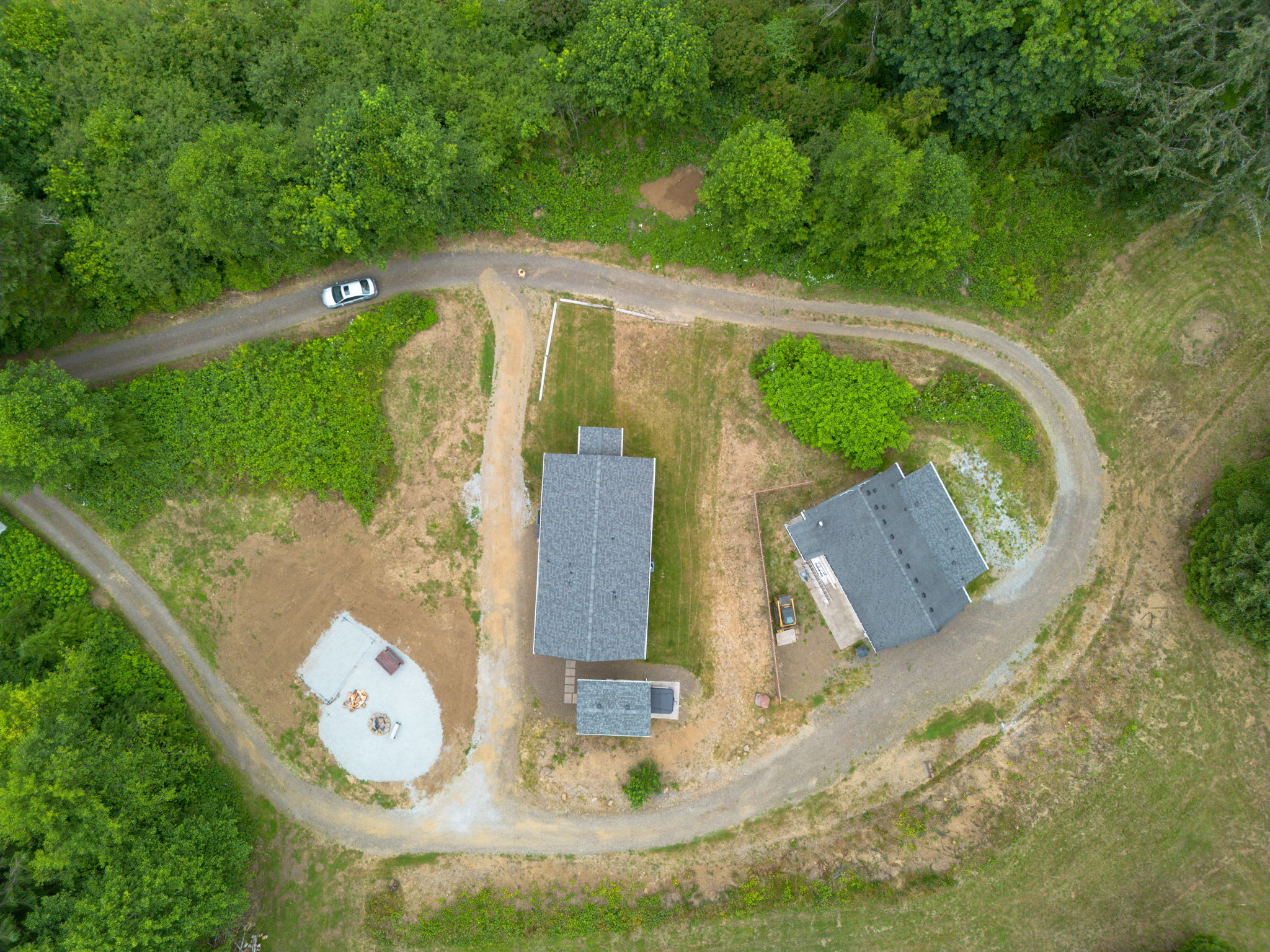 A top-down aerial view shows a winding gravel driveway encircling two houses and a fire pit area, all set within a clearing surrounded by dense green trees. A white car is parked on the driveway.