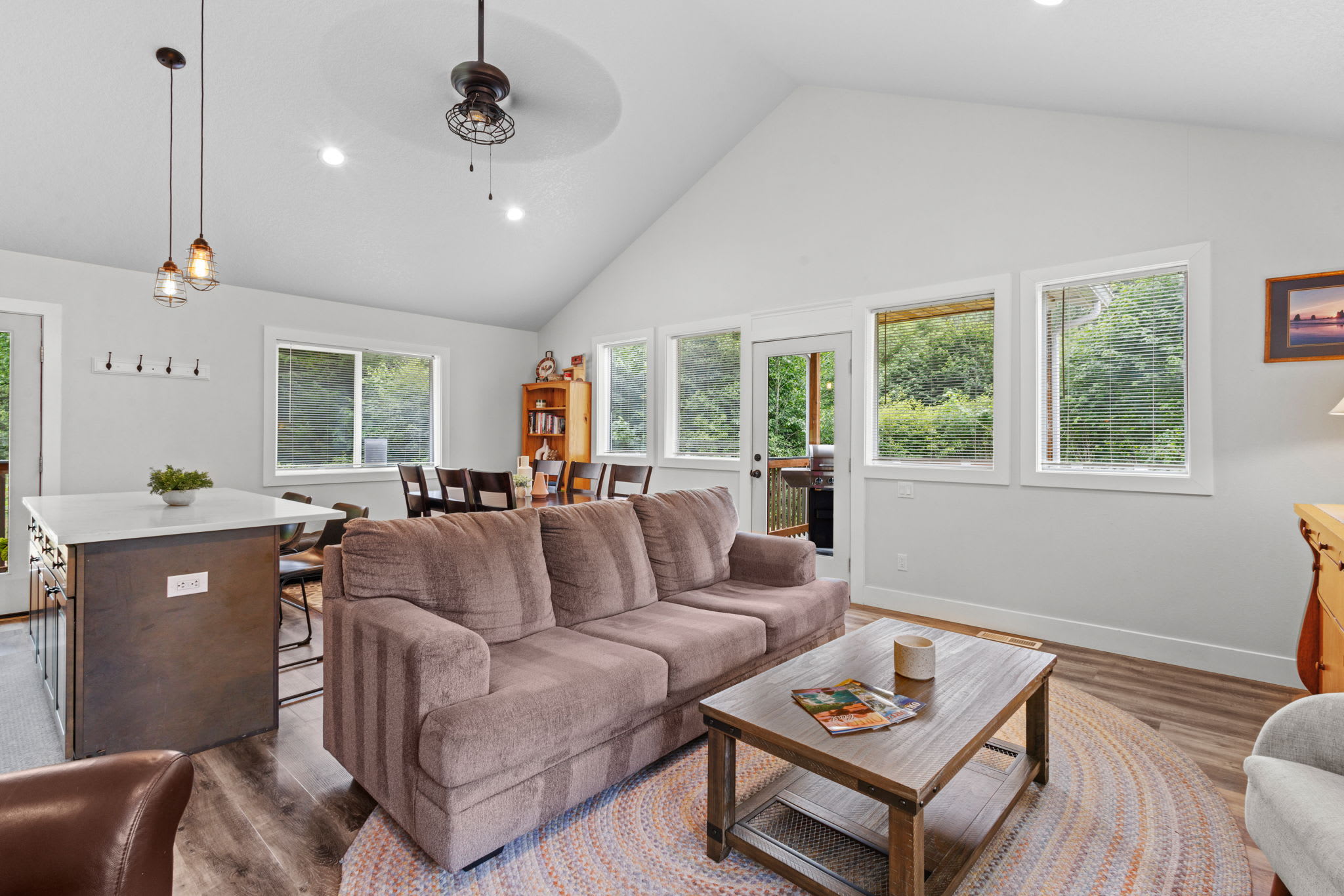 Cozy living room featuring a large brown sofa and a wooden coffee table on a round patterned rug, with the dining area and kitchen visible in the background, under a vaulted ceiling with a ceiling fan.