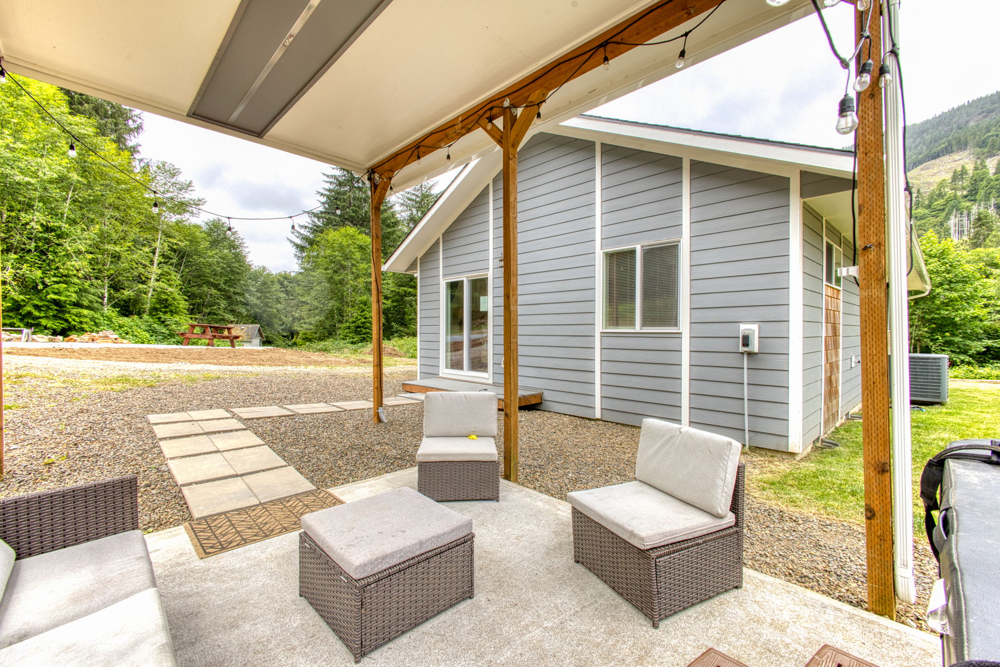A covered outdoor patio with string lights features wicker seating with light cushions, a concrete slab, and stepping stones leading to a gravel area. The light gray house with a vaulted ceiling is visible in the background, surrounded by green trees.