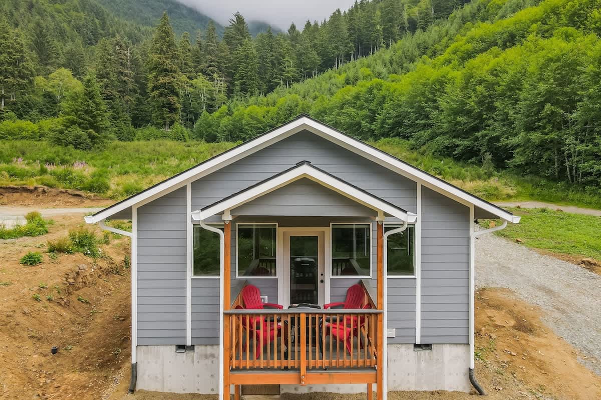 The front view of a gray house with white trim, featuring a small wooden porch with two red Adirondack chairs. The house is set on a slight incline with a gravel path leading up to it, surrounded by green hills and dense forest.