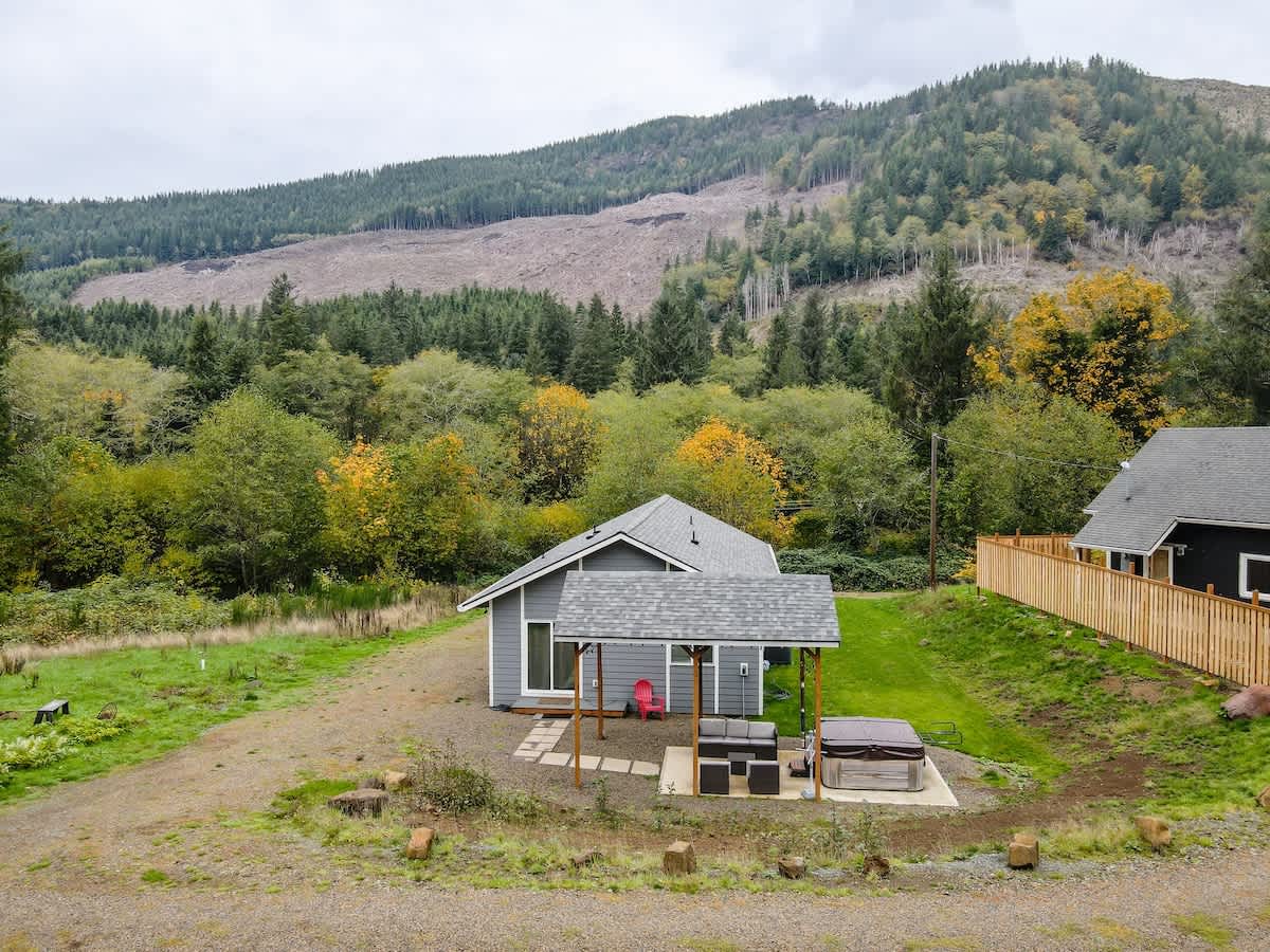 An exterior view of a gray house with a covered patio featuring outdoor seating and a hot tub, set against a backdrop of a forested mountain with patches of autumn colors. A gravel area surrounds the house, and a dark house is visible in the distance.