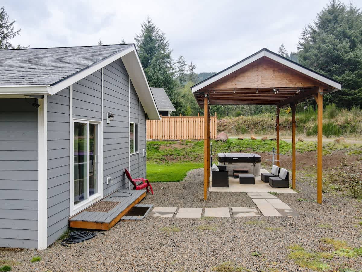 A close-up view of the side of a gray house with a sliding glass door, leading to a gravel patio with a covered hot tub and outdoor seating area. A wooden fence and lush green hills are visible in the background.