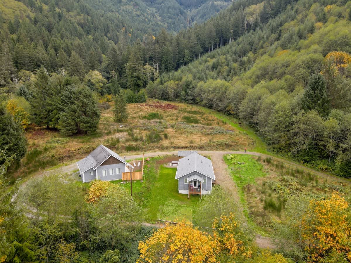 An aerial view showing two houses, one gray and one dark, nestled in a clearing within a vast, lush green valley surrounded by dense forests. A gravel path connects the properties, and a wooden deck is visible near the dark house.