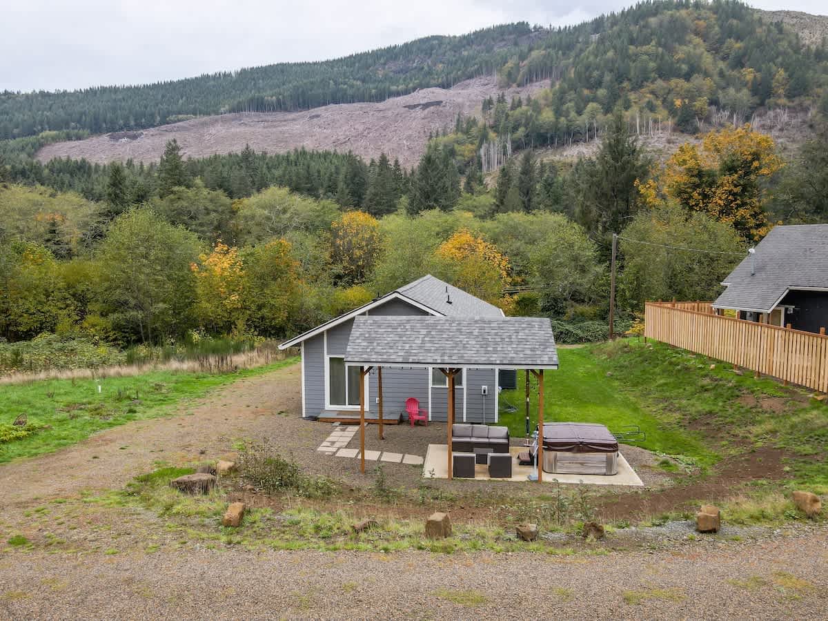 An exterior shot of a gray house with a covered patio, outdoor seating, and a hot tub, surrounded by a gravel yard and green grass. A large forested mountain with some cleared areas rises in the background, showing hints of autumn foliage.