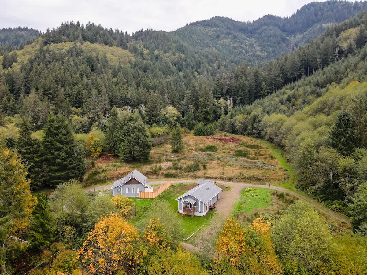 An aerial view shows two light gray houses nestled in a valley surrounded by dense green forests and rolling hills, with a dirt road winding through the property. The autumn trees in the foreground add a touch of yellow and orange.