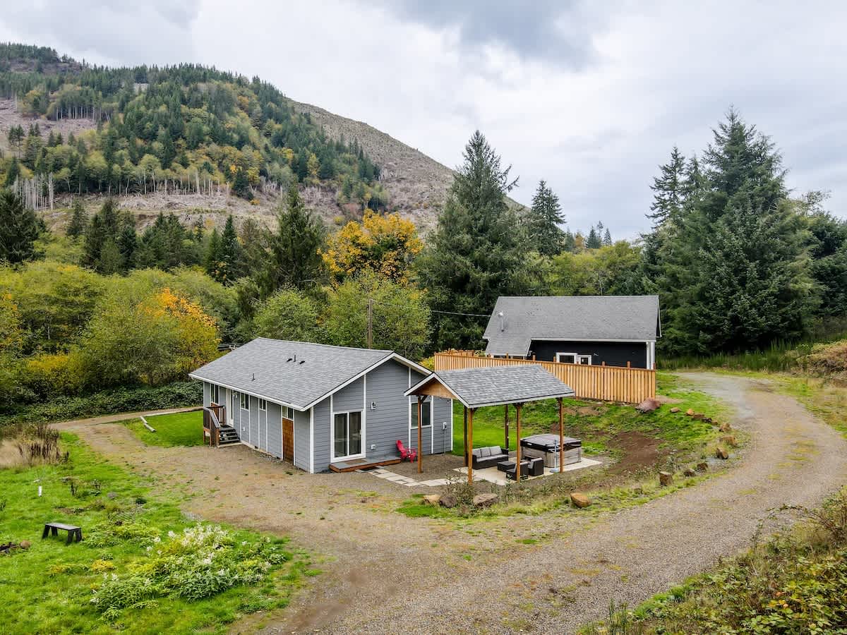 An aerial view showing two houses, one gray and one dark, nestled in a valley surrounded by dense green and autumnal forests with a mountain in the background. A gravel path connects the houses, and a covered hot tub area is visible near the gray house.