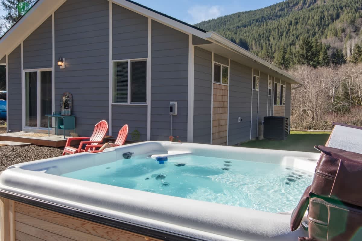 A sparkling clean hot tub sits on a wooden deck next to a modern gray house with white trim, surrounded by green grass and distant forested mountains under a bright sky.