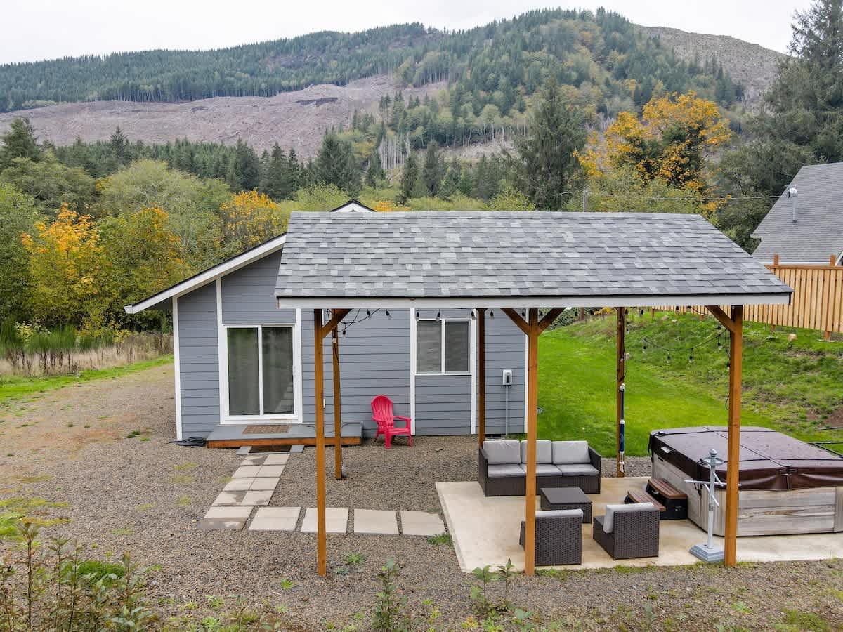 A ground-level view of the gray house's side, featuring a covered patio with outdoor seating and a hot tub, surrounded by gravel and a small patch of grass. A forested mountain with autumn colors provides a scenic backdrop.