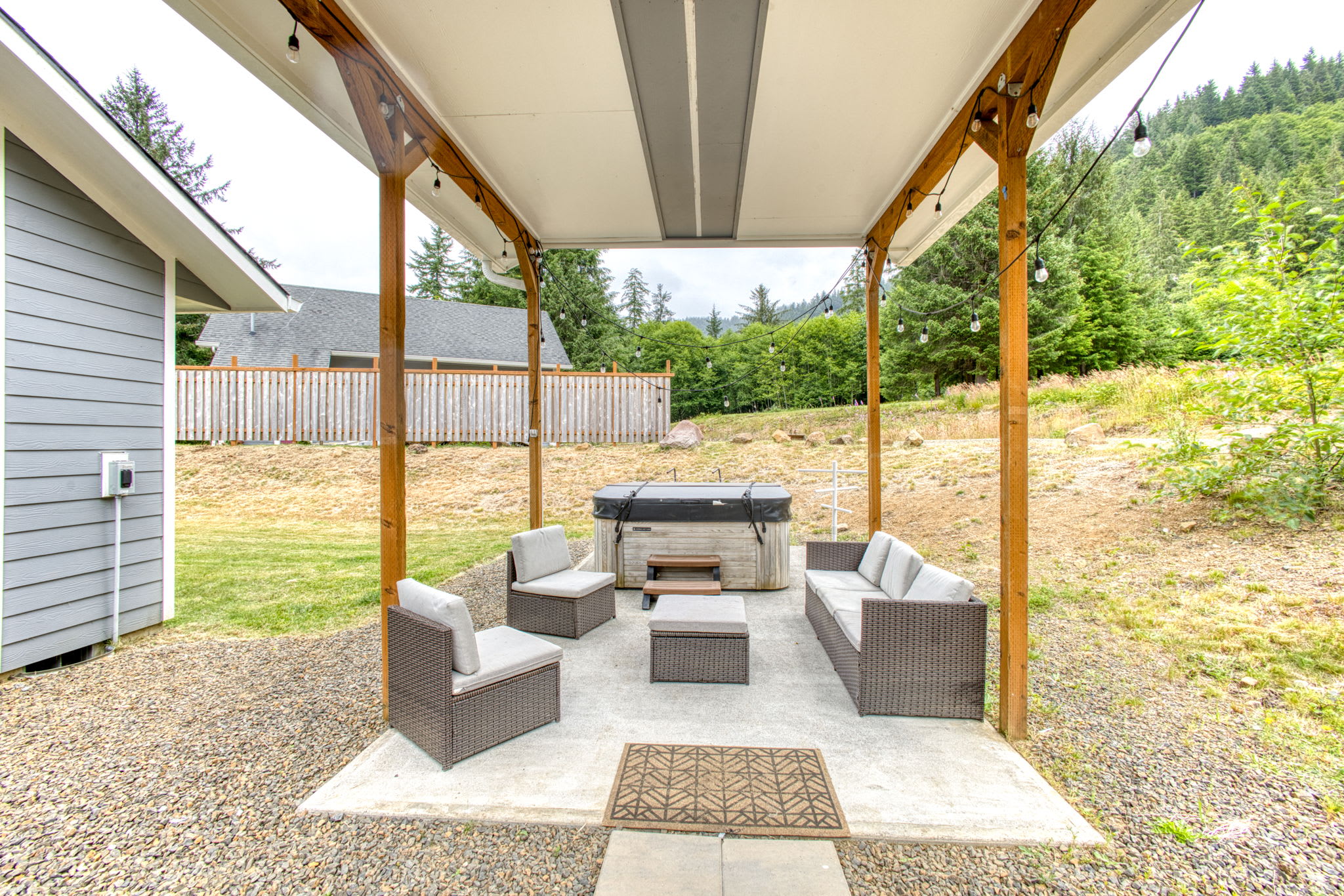 A covered outdoor patio area featuring a hot tub, comfortable wicker outdoor seating with light cushions, and string lights overhead. The area is set on a concrete slab surrounded by gravel and backed by a grassy hill and lush trees.