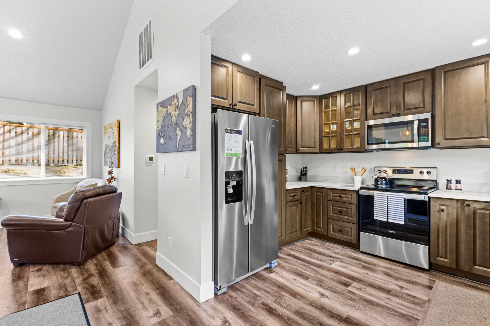 Spacious open-concept living and dining area with vaulted ceilings, featuring a dark wood dining table with six chairs, a comfortable sofa, and large windows offering natural light and views of the surrounding greenery.