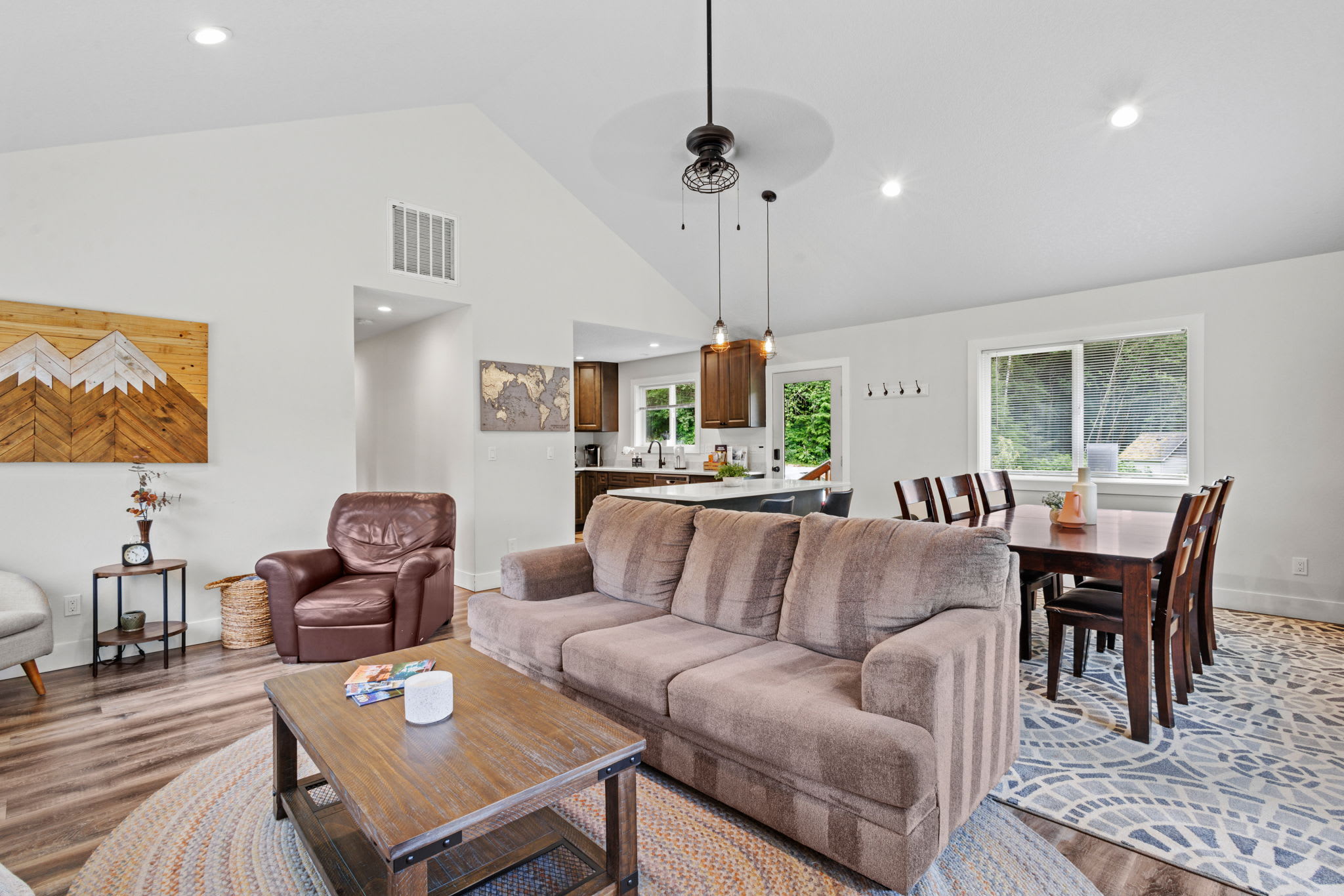 Spacious open-concept living room with a brown sofa, wooden coffee table, and a brown leather armchair, leading into the dining and kitchen areas, featuring hardwood floors and a vaulted ceiling.