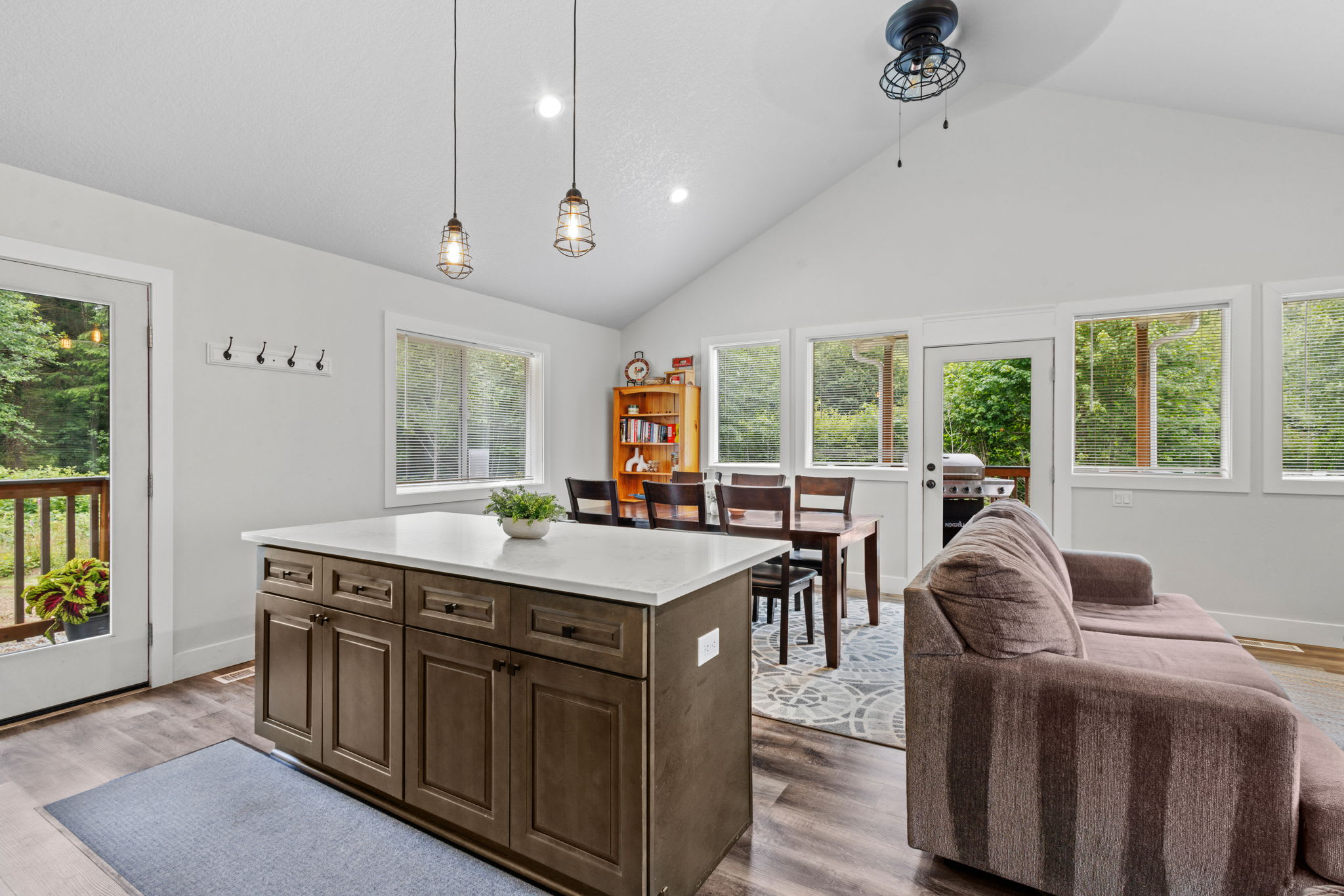 Open-concept living space featuring a dark brown kitchen island with a white countertop, a matching dining table with six chairs, and a glimpse of a brown sofa, all under a vaulted ceiling with pendant lights.