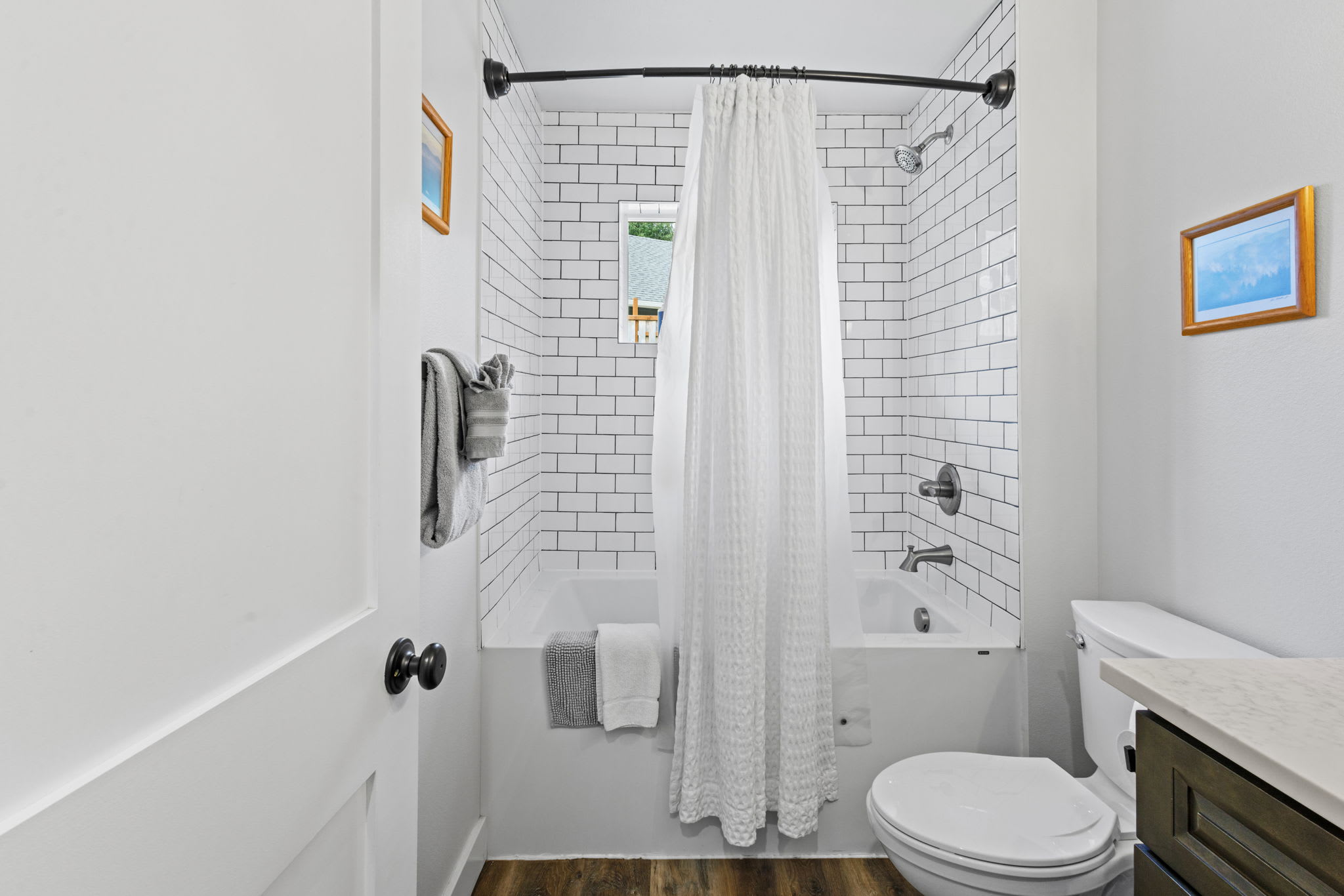 A clean bathroom with a white bathtub surrounded by white subway tiles with dark grout, a white waffle-textured shower curtain, and a toilet positioned next to the tub. A small framed picture hangs on the wall.