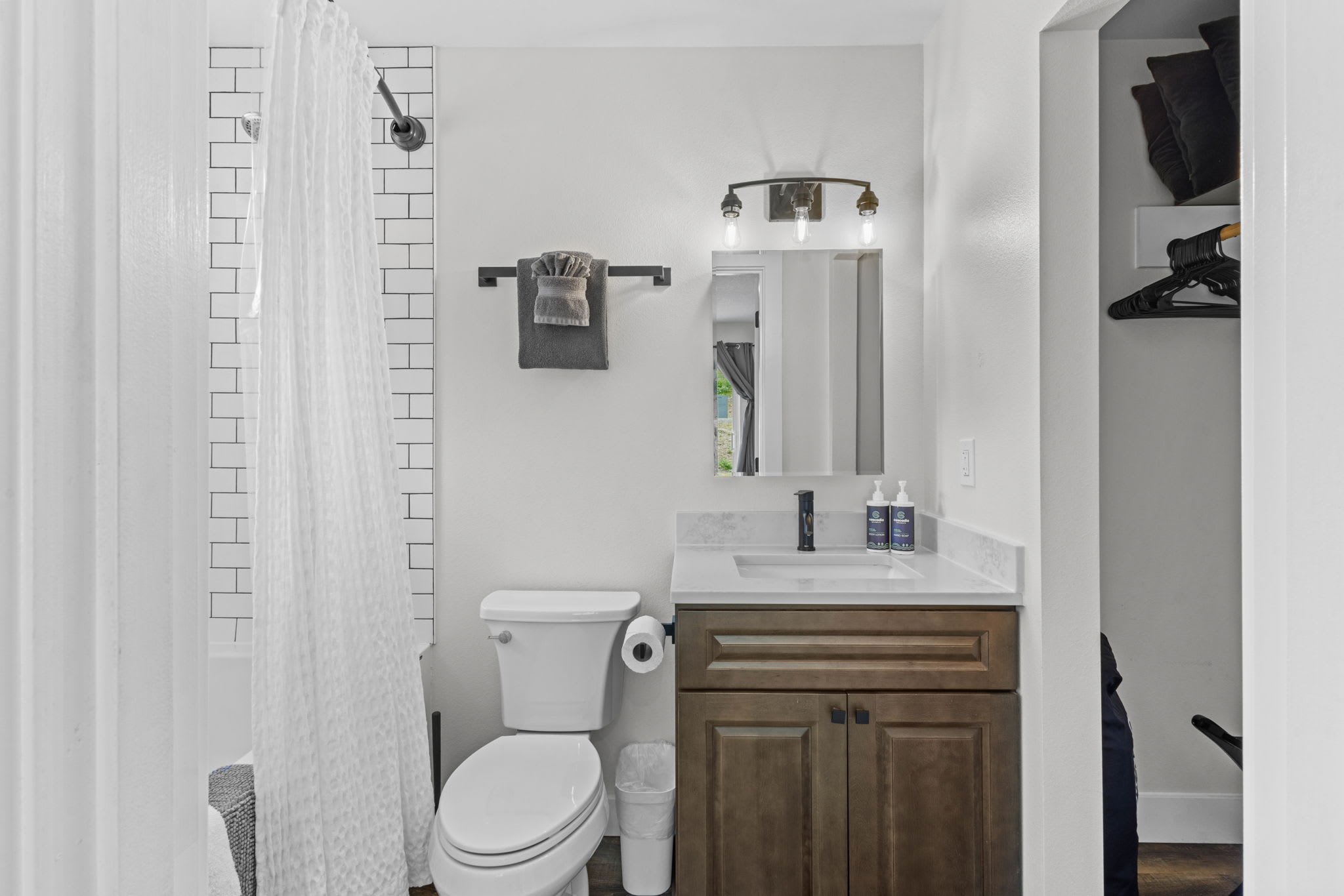 A modern bathroom with a dark wood vanity, white quartz countertop, rectangular sink, black faucet, and a toilet next to a white trash can. A mirror with industrial-style lighting hangs above the vanity.