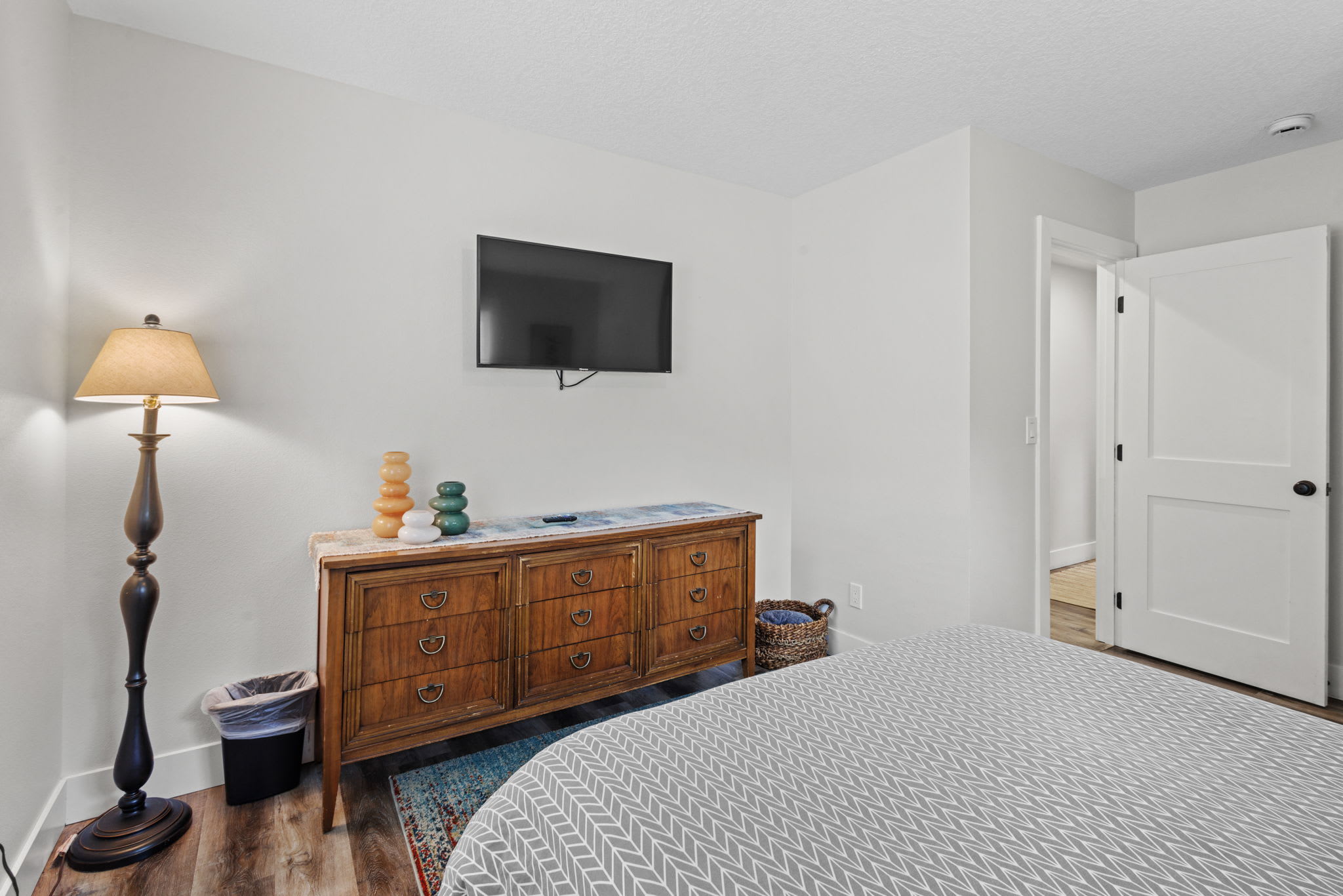 A bedroom view showing a vintage wooden dresser with a light-colored marble top, a wall-mounted flat-screen TV, and a tall floor lamp with a beige shade. A colorful area rug is partially visible on the dark wood-look flooring.