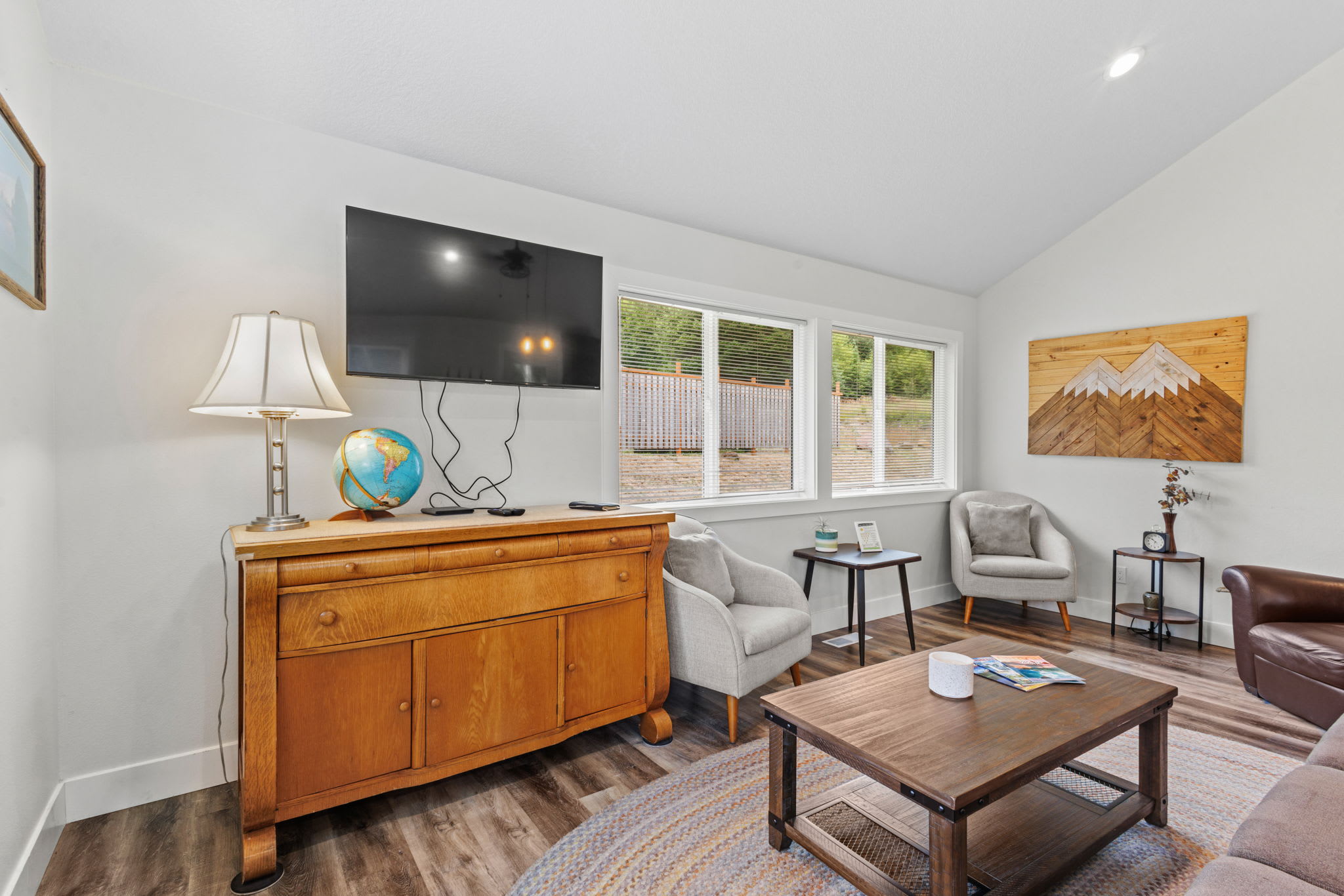 Living room area featuring a wall-mounted flat-screen TV above a vintage wooden credenza, two light grey armchairs, and a wooden coffee table, with large windows overlooking the outdoors.