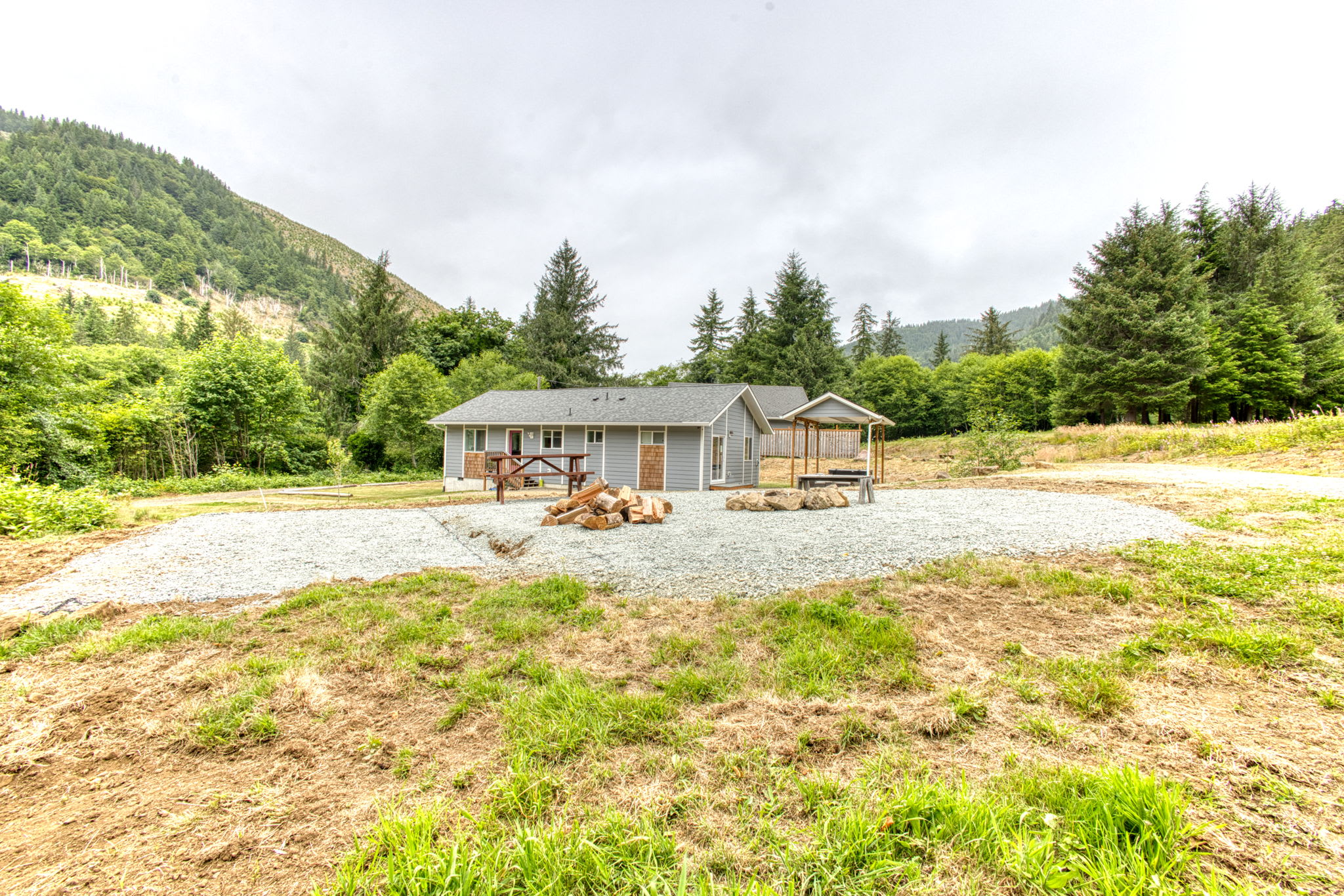 A wide shot of the property shows a large gravel area with a stone fire pit, wooden benches, and a picnic table, with stacked firewood nearby. Two light gray houses are visible in the background, surrounded by green trees and a distant mountain.