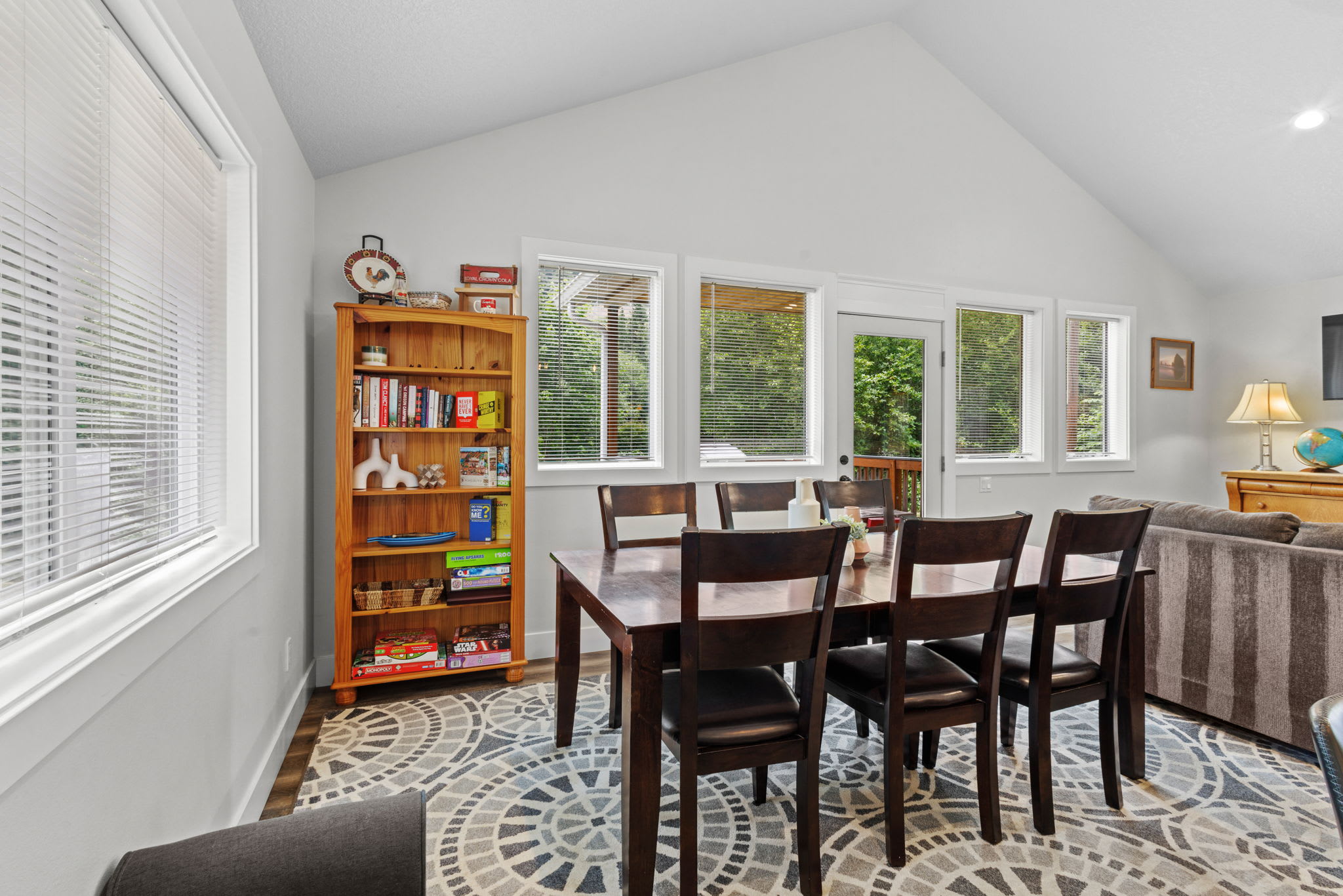 Dining area featuring a dark wood table with six chairs on a patterned rug, next to a wooden bookshelf filled with books and games, with multiple windows offering views of the surrounding trees.