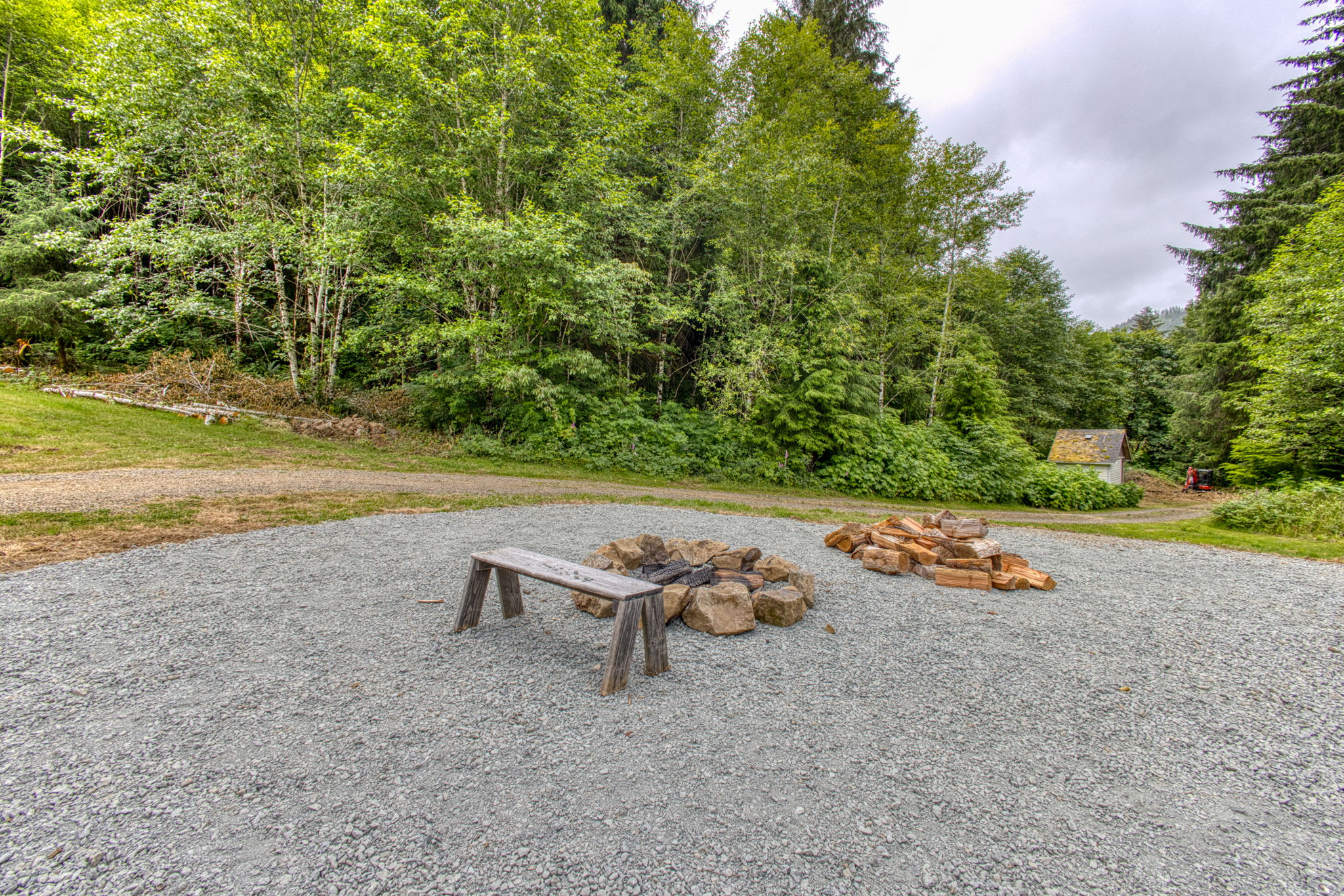 An outdoor fire pit area set on a gravel surface, featuring a stone-ringed fire pit, a rustic wooden bench, and a stack of firewood. The area is surrounded by green grass and dense forest in the background.
