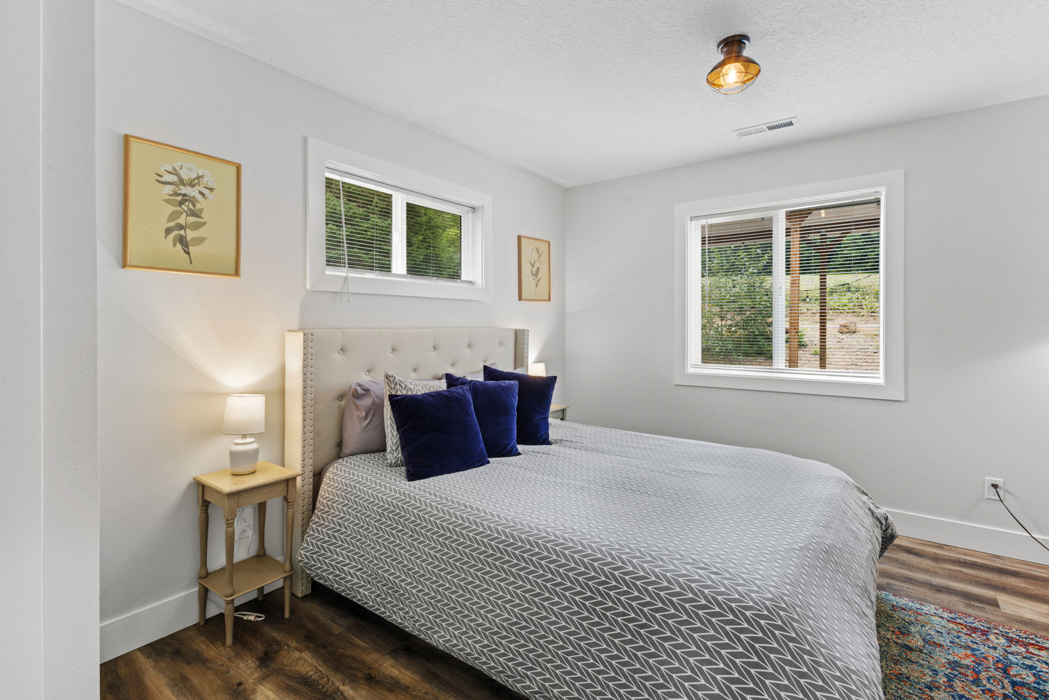 A bright bedroom with a queen-sized bed, light gray tufted headboard, and two windows with blinds providing natural light. The room has light walls, dark wood-look flooring, and two framed botanical prints on the walls.