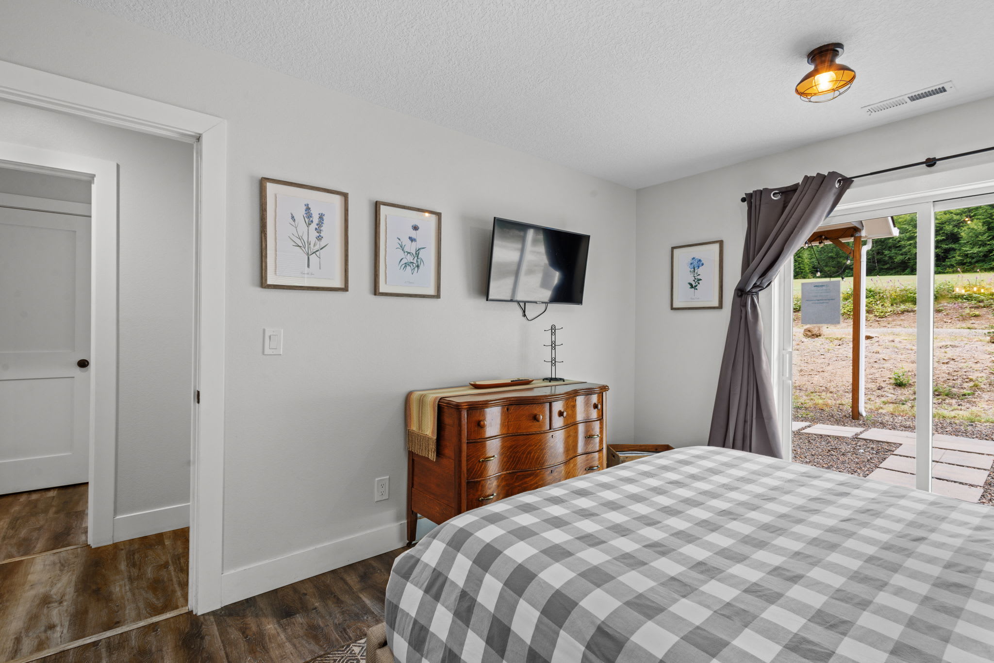 Bedroom with a bed featuring a grey and white checkered duvet, a wooden dresser with a TV mounted above it, and a glass sliding door leading to an outdoor patio area.