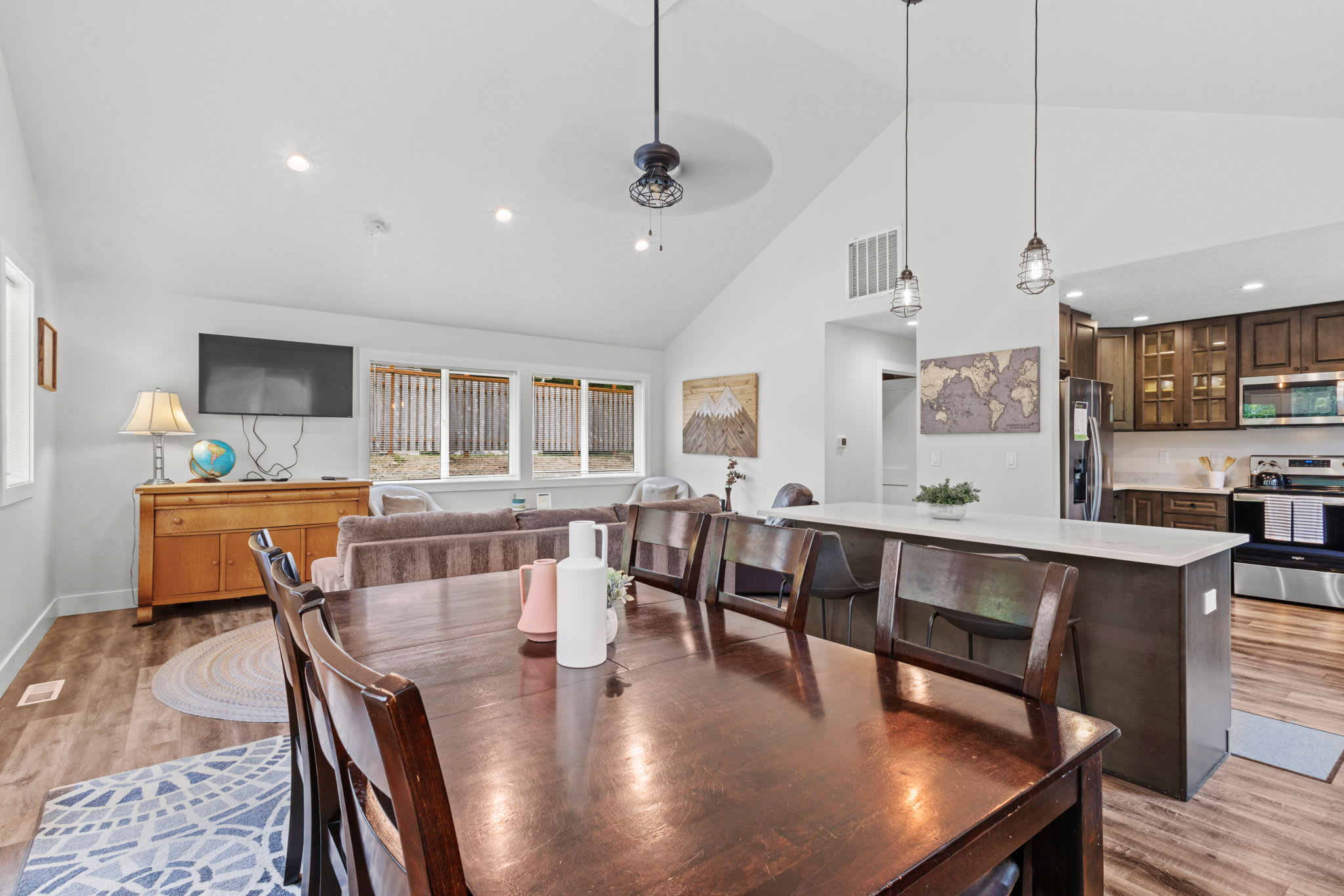 View of the dining area with a dark wood table and chairs, looking towards the kitchen island with bar seating and the living room beyond, all within an open-concept space with vaulted ceilings.