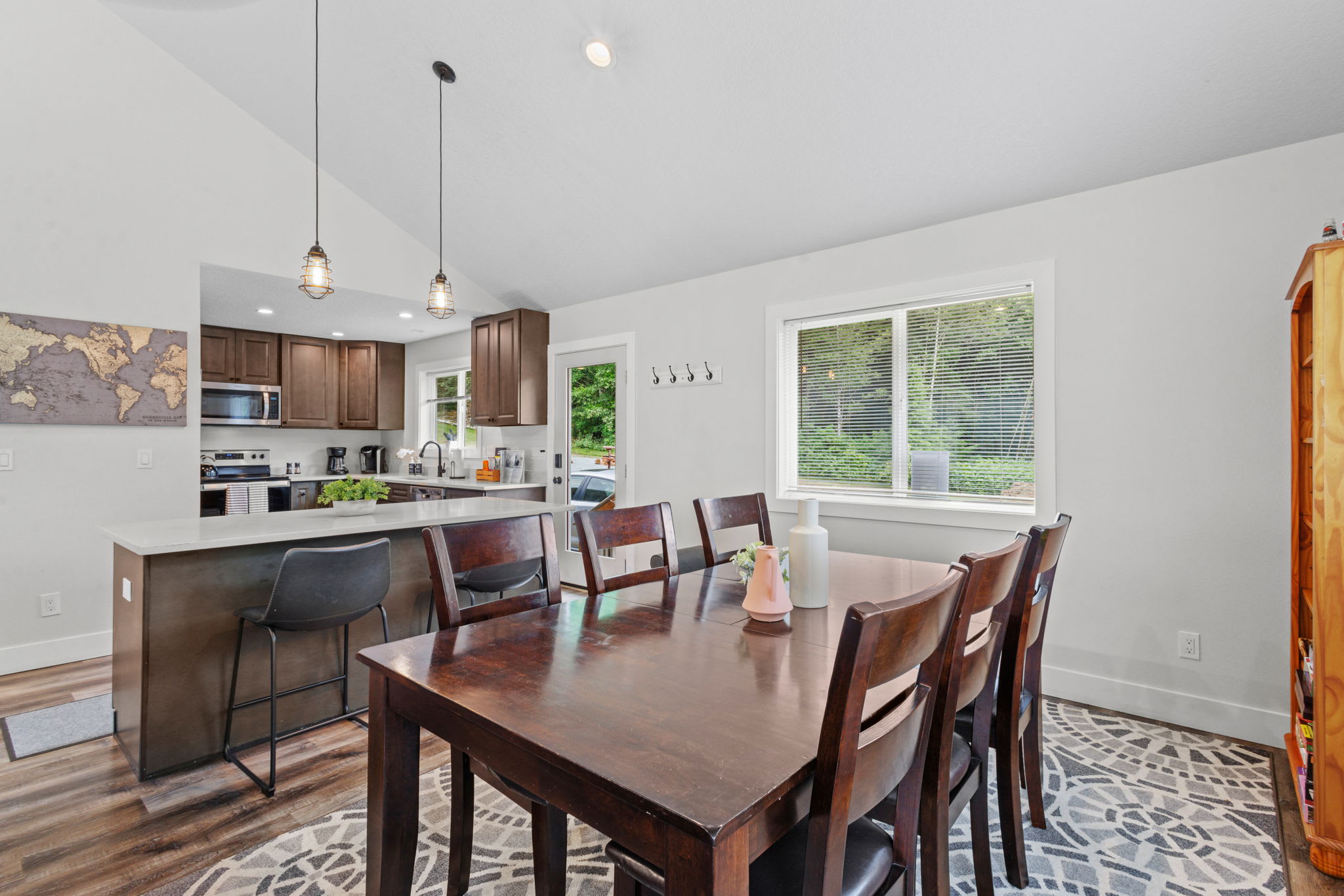 Modern kitchen with dark wood cabinets, stainless steel appliances, a large island with white countertop and bar stools, and an adjacent dining area with a dark wood table and chairs, all under a vaulted ceiling.