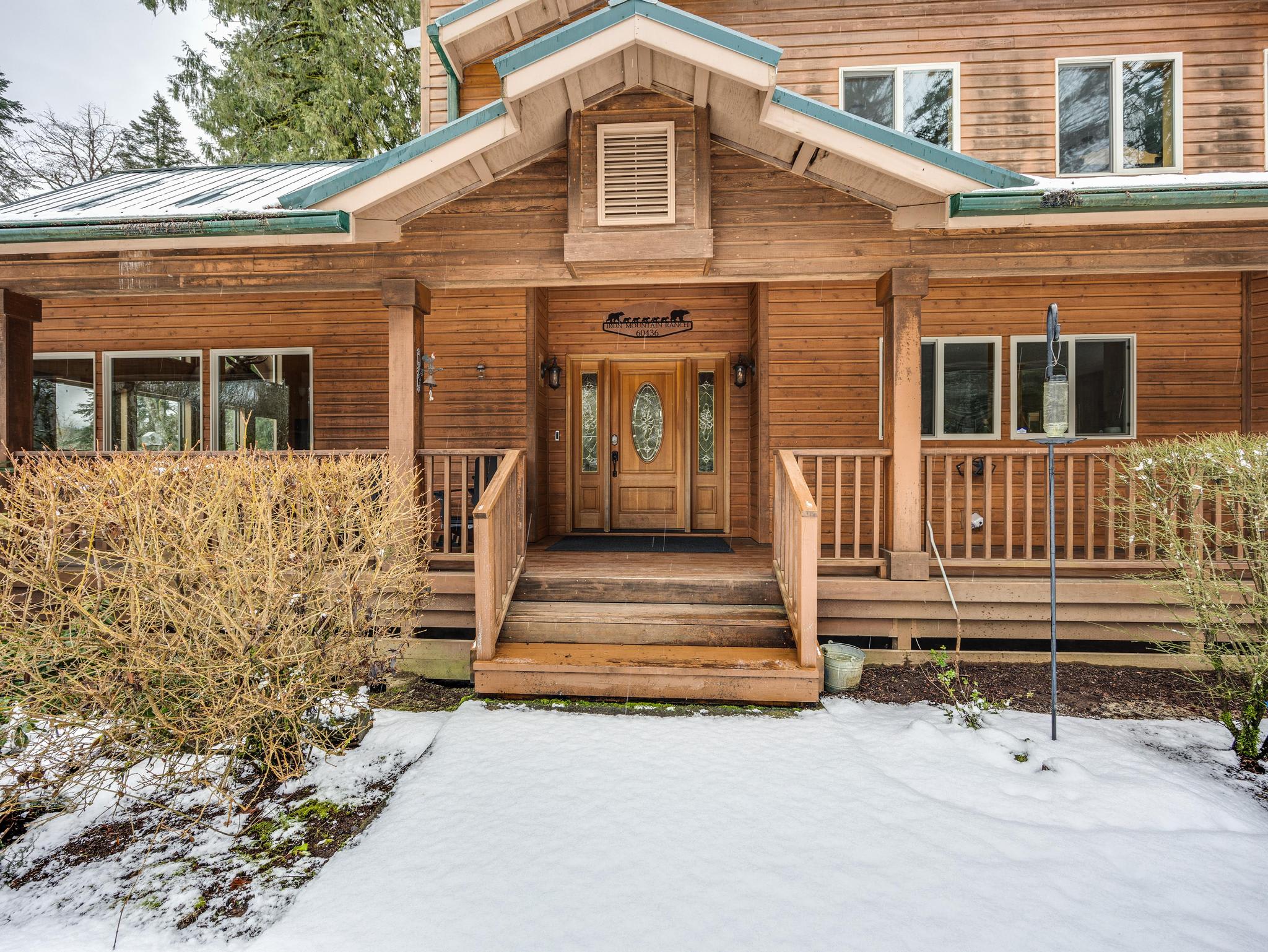 Inviting front entrance of the wooden lodge, featuring a covered porch with wooden railings and steps leading to a decorative glass-paneled door, with a light dusting of snow on the ground.