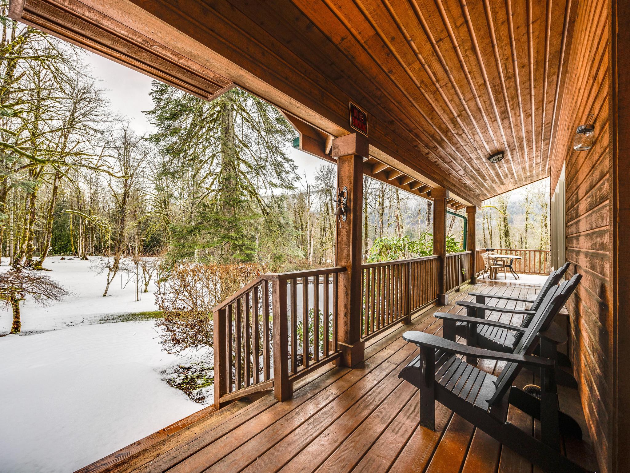 A covered wooden porch with several dark Adirondack chairs offers a view of a snow-covered landscape and bare trees, showcasing the property's winter appeal.