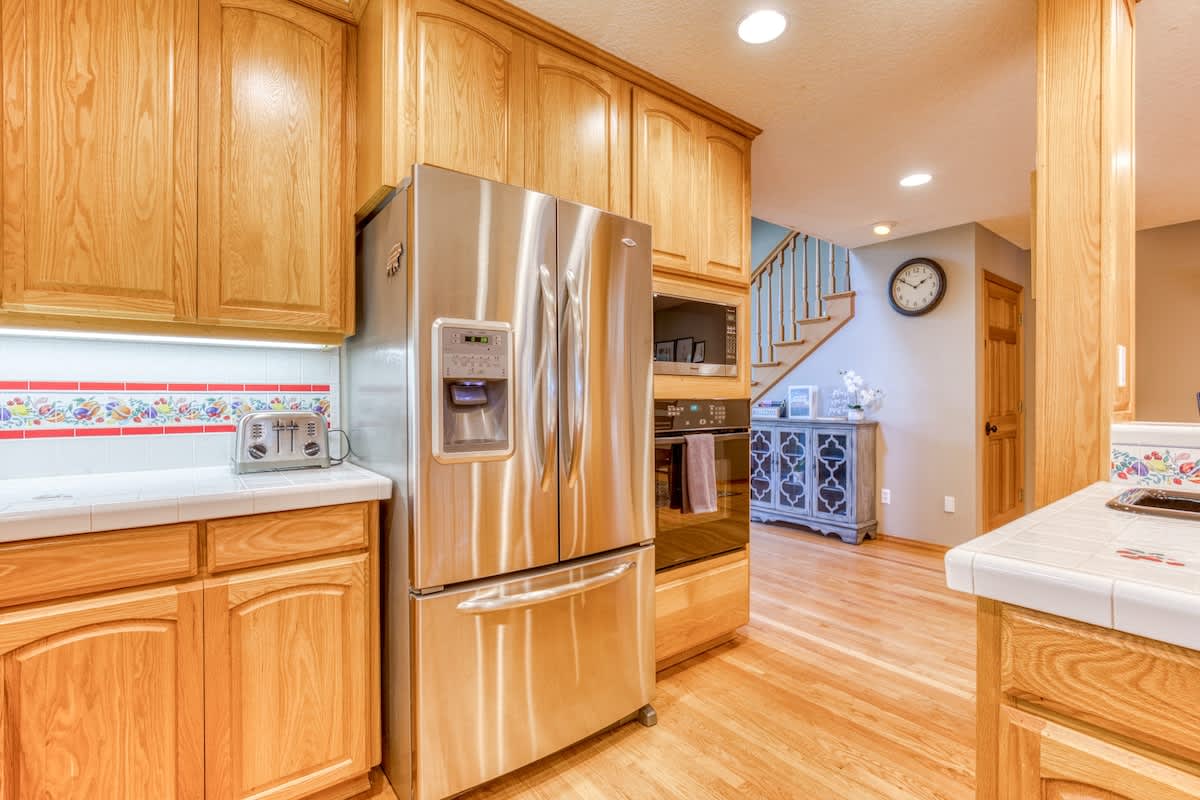 Modern kitchen corner showcasing a stainless steel French door refrigerator and a built-in oven and microwave. Light wood cabinetry and white tiled countertops extend around the space, with a decorative tiled backsplash.