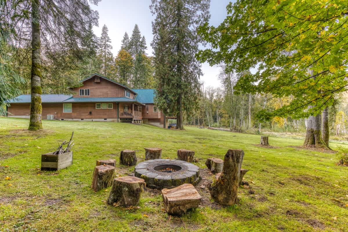 A stone fire pit is surrounded by rustic tree stump seating in a large, grassy backyard, with a wooden house and dense forest visible in the background under a soft, overcast sky.
