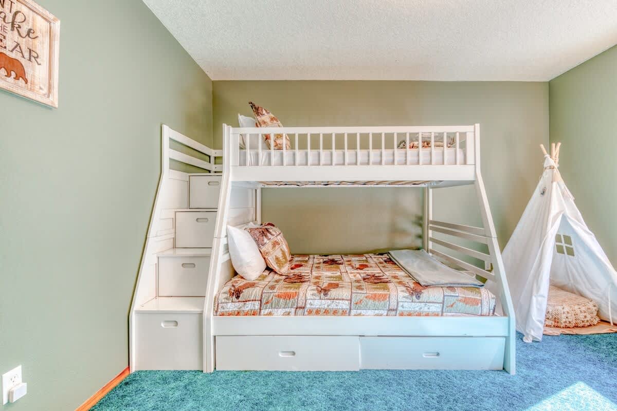 A children's bedroom showcasing white wooden bunk beds with built-in storage stairs, a lower full bed, and an upper twin, complemented by a white teepee and a vibrant teal carpet.